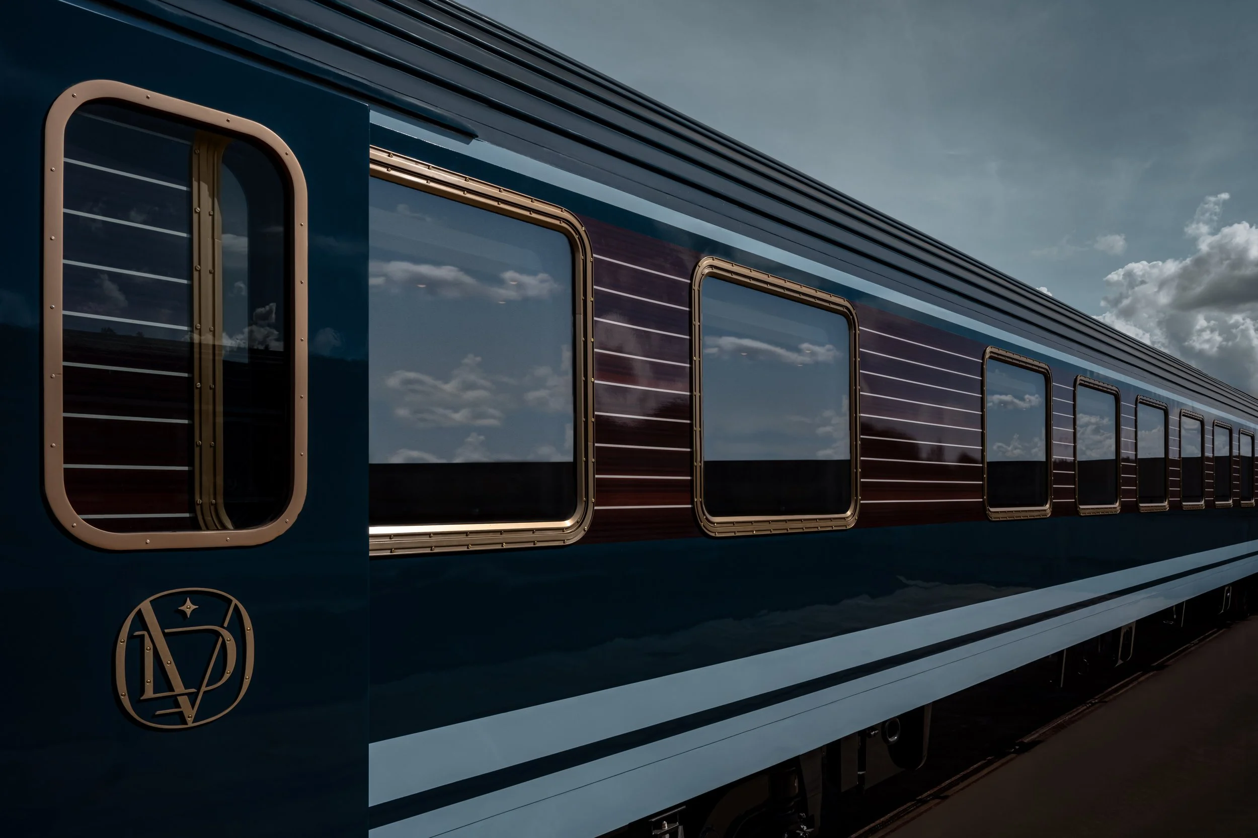 Close-up view of a train with blue and black horizontal stripes and reflective windows reflecting clouds and sky.