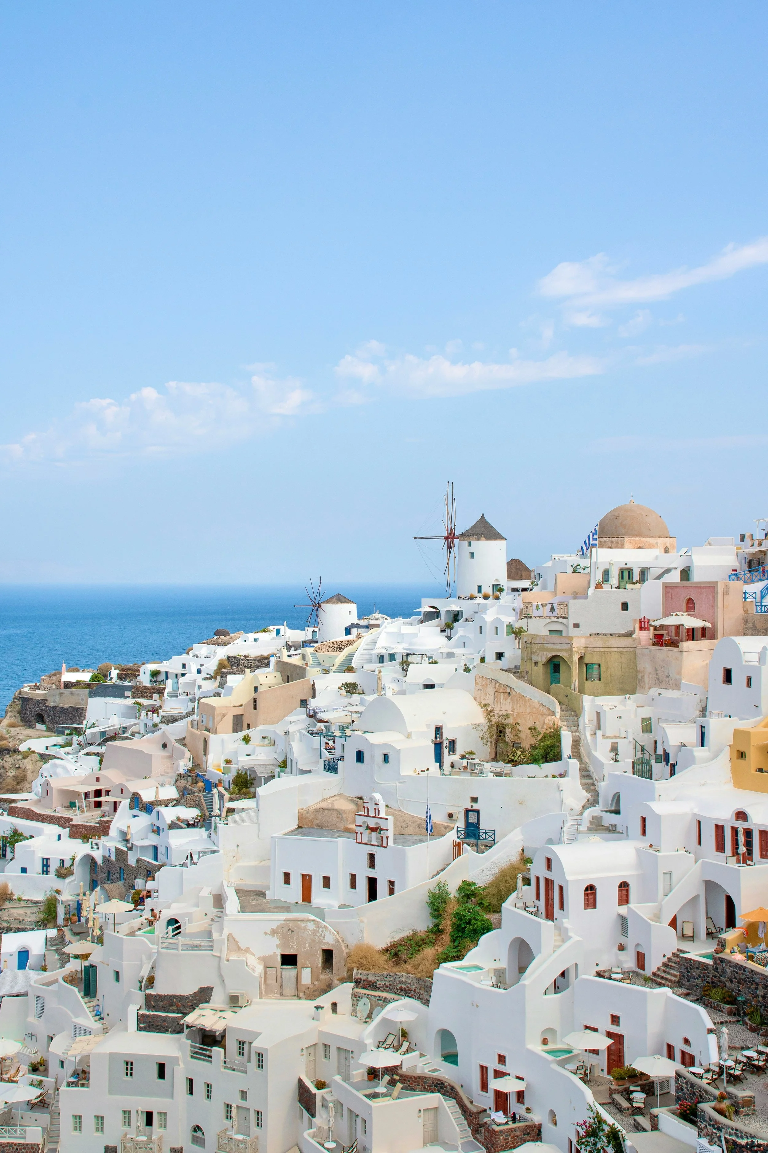 White buildings with blue accents and windmills on a hill overlooking the sea, typical of Santorini, Greece, under a partly cloudy sky.
