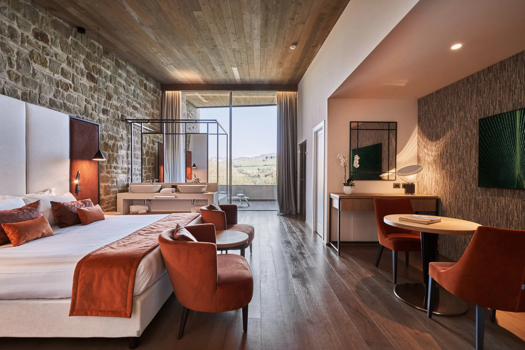 Luxurious hotel room with a large bed adorned with rust-colored pillows and a matching throw blanket, a seating area with velvet chairs, and a balcony with scenic view. The room features exposed stone and wood-paneled walls, modern lighting, a vanity with sinks, and stylish decor with plants and artwork.