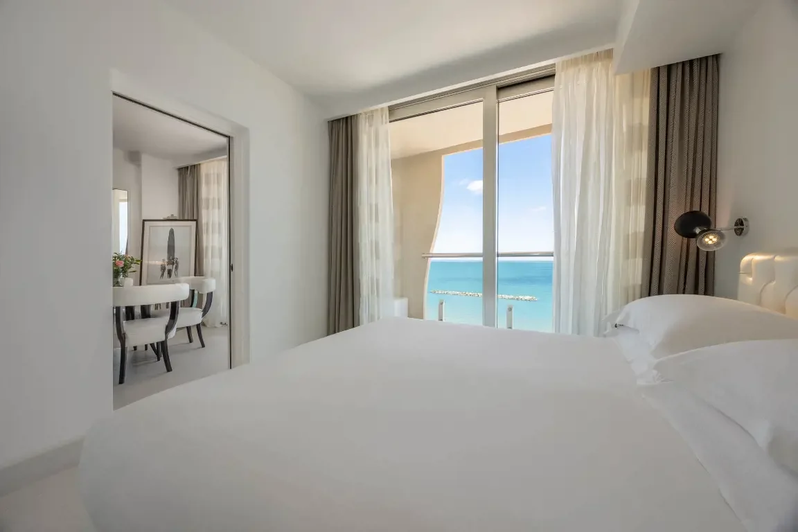 A bright hotel room with a large bed, ocean view through glass sliding doors, and a balcony with a railing.