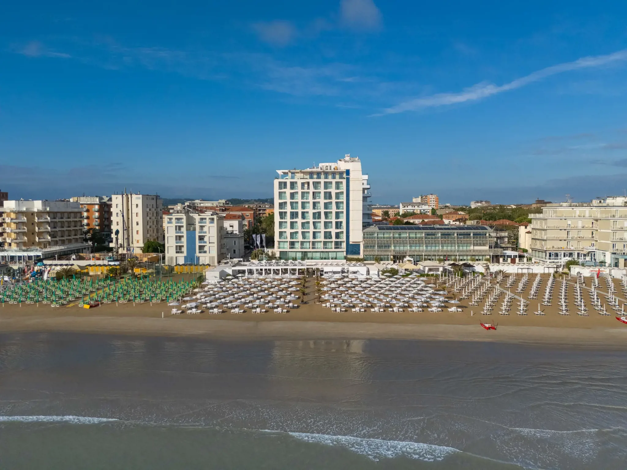 A cityscape with tall modern buildings and a beach in the foreground, featuring organized rows of umbrellas and lounge chairs by the water under a clear blue sky.