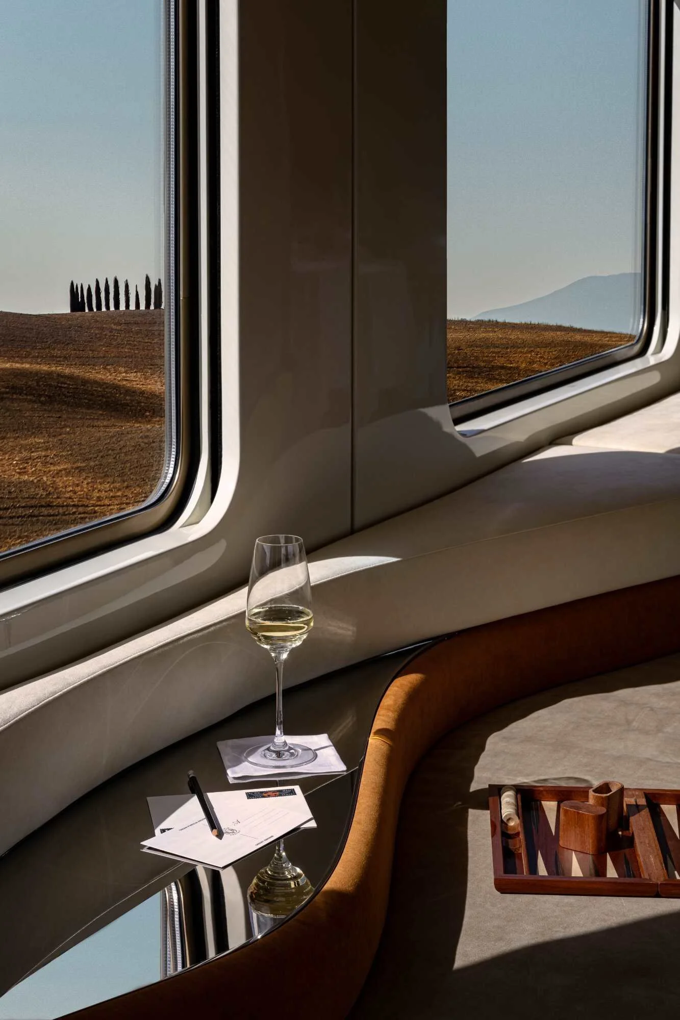 A room with a curved window ledge, a glass of white wine on a stand, and a tray with cigars, with a landscape view of hills and a clear sky outside.
