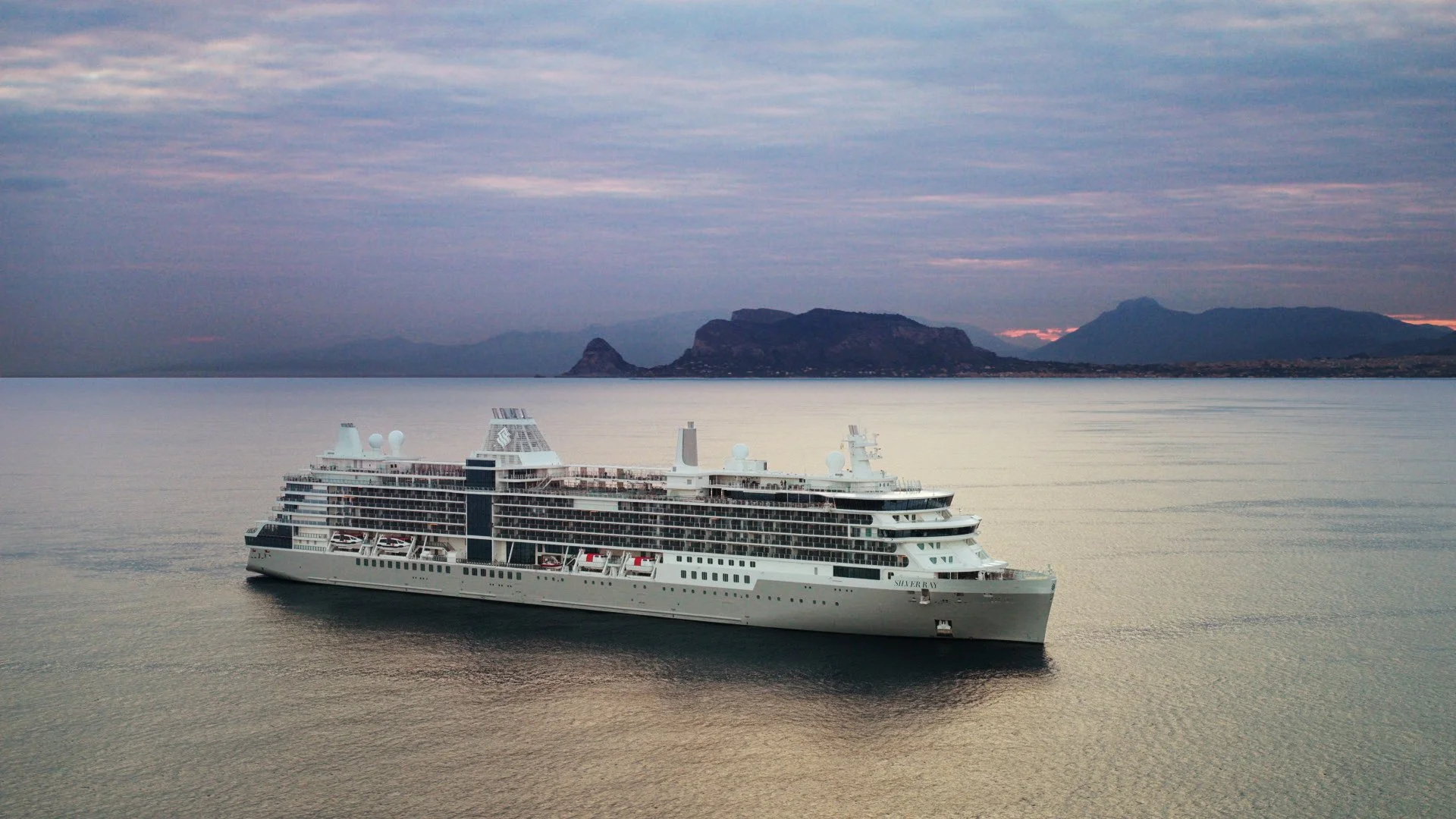 A large white cruise ship sailing on calm water with mountainous landscape in the background during dusk or dawn.