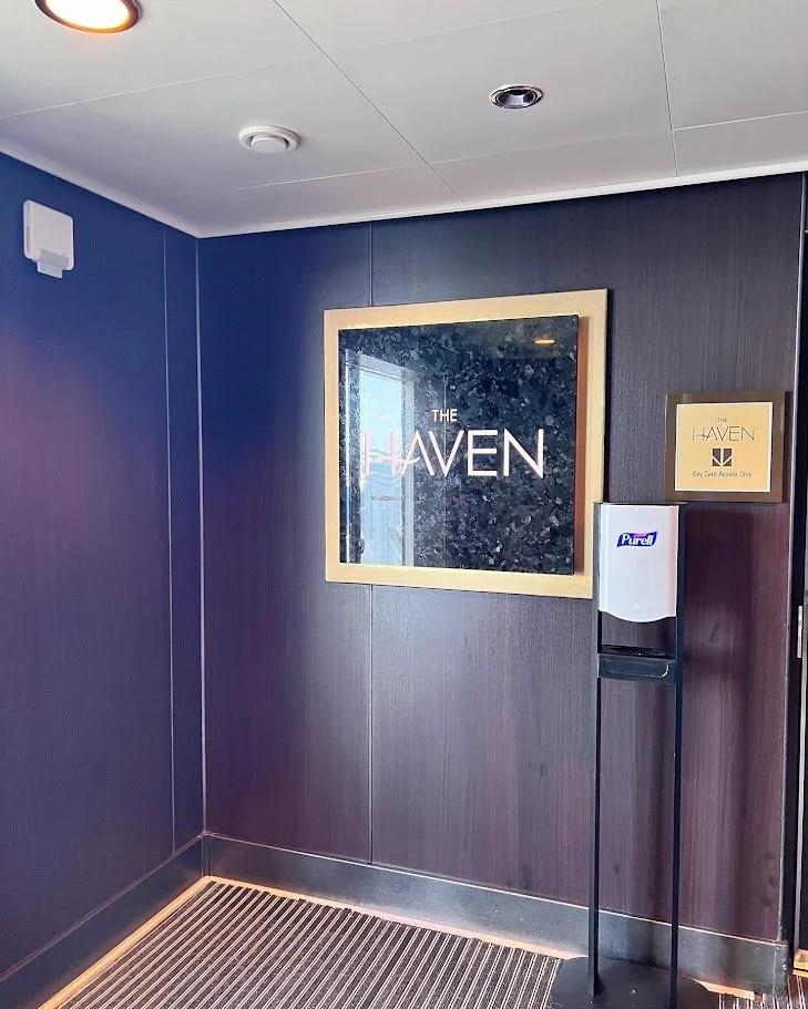 Interior of a building with a sign that says 'The Haven', and a hand sanitizer dispenser nearby. The wall is dark wood, and there is a window reflected in the sign.