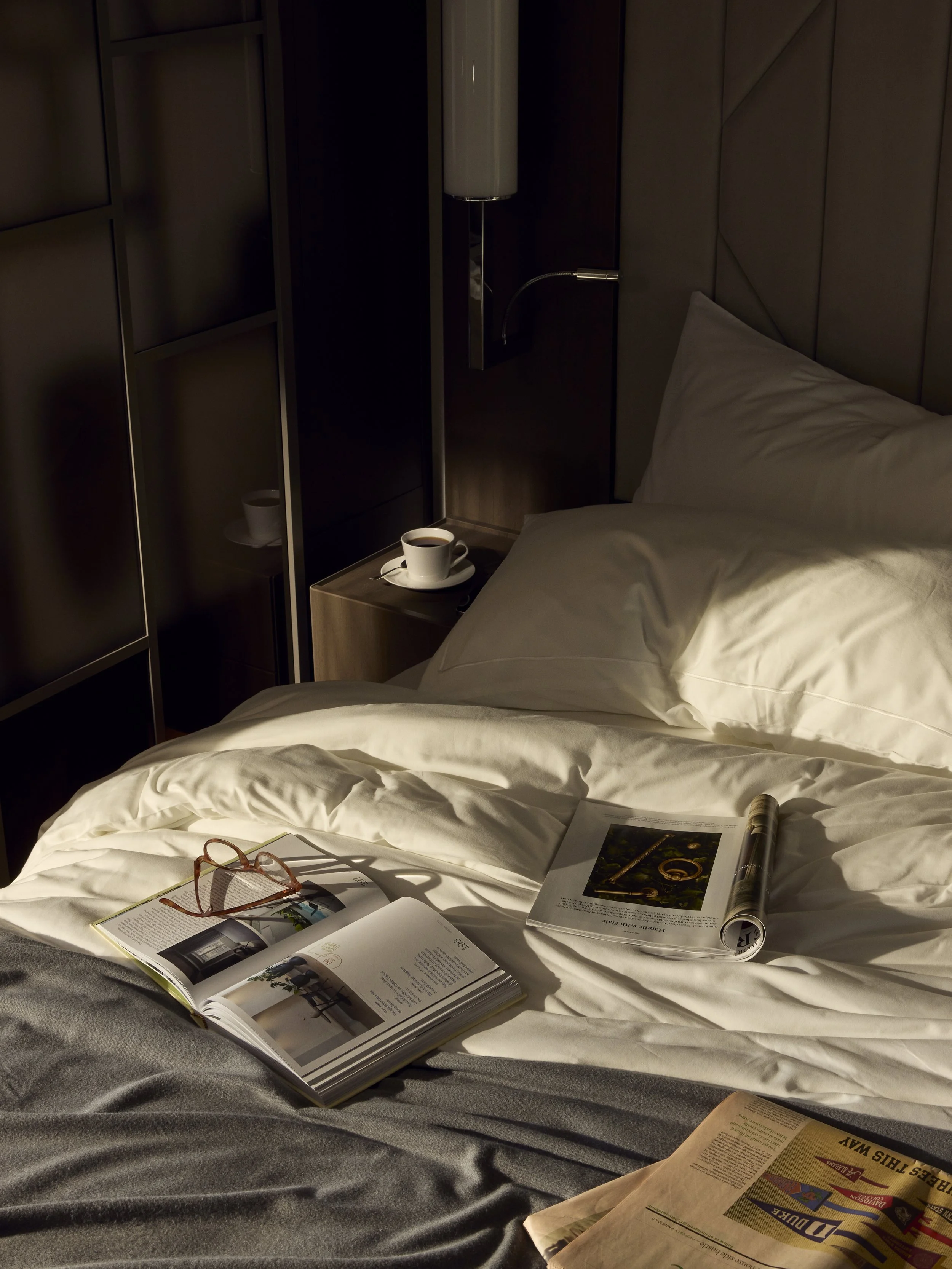 A bed with open magazines and papers, a pair of glasses, and a cup of coffee on the nightstand. The bed has white sheets and a pillow, with a dark wall and some furniture in the background.