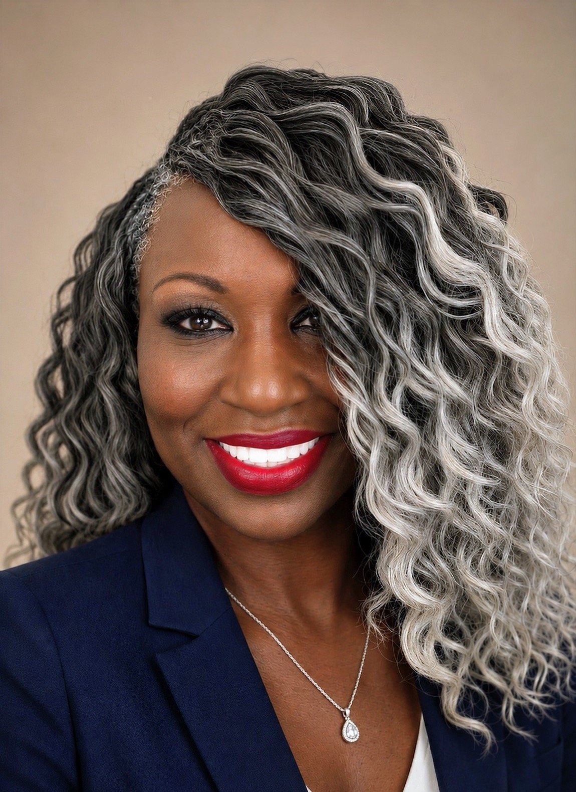 A woman with curly, gray and black hair styled to the side, smiling with red lipstick, wearing a dark blazer, a white top, a silver necklace with a teardrop pendant, and makeup.