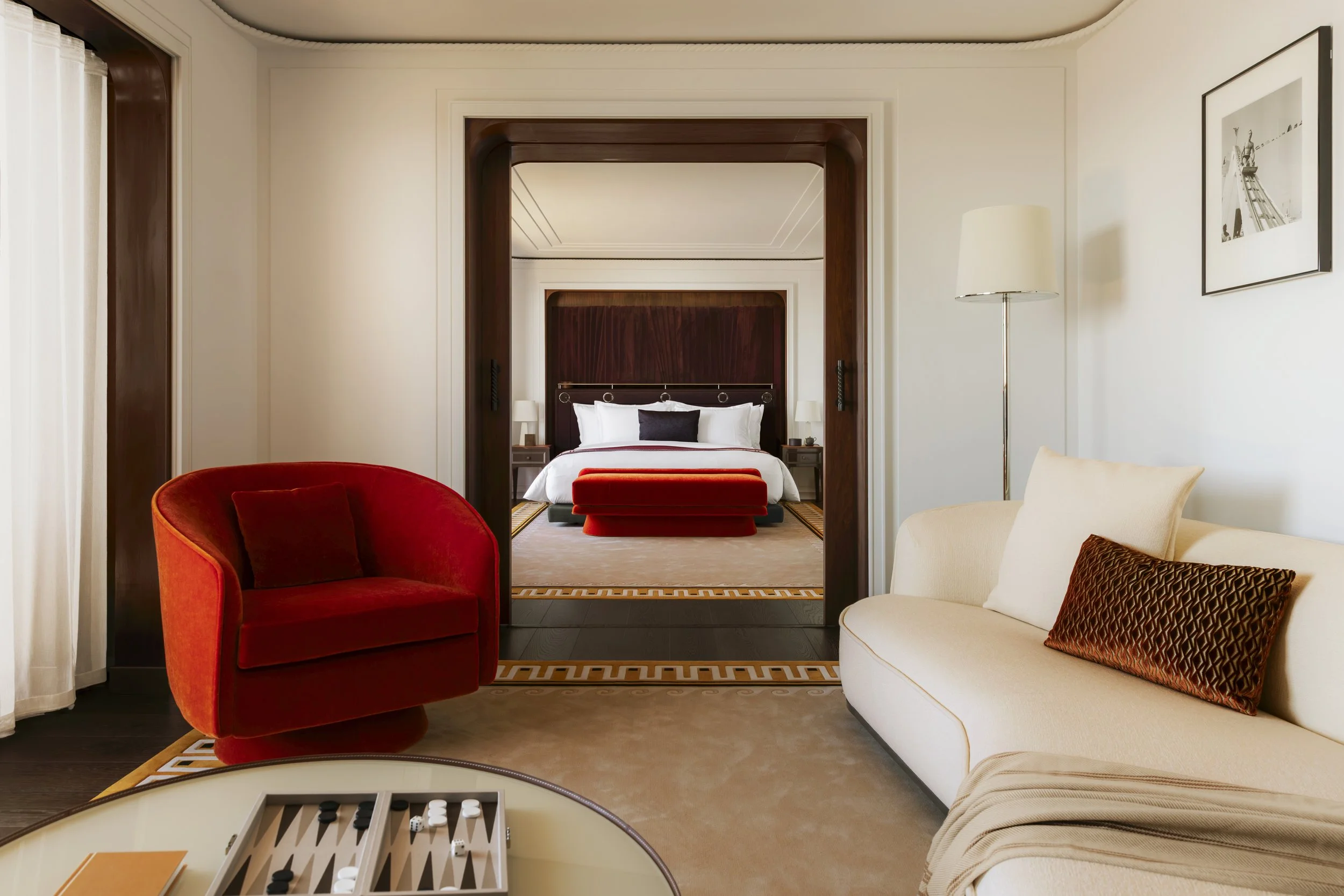 View through doorway into a bedroom with a bed, two bedside tables, and lamps. In the foreground, a red upholstered armchair and cream-colored sofa with decorative pillows. A framed black and white photograph on the wall, and a floor lamp.
