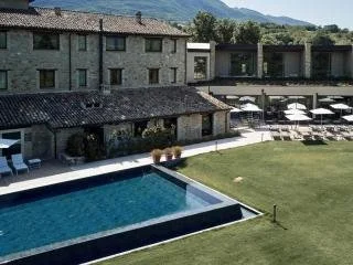 Outdoor view of a modern hotel or apartment complex with a large swimming pool, lounge chairs, and umbrellas on a grassy area, with mountains in the background.