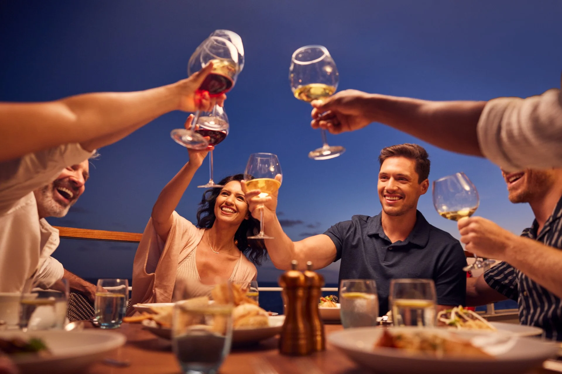A group of friends enjoying a toast with wine glasses at an outdoor dinner during the evening, smiling and celebrating.