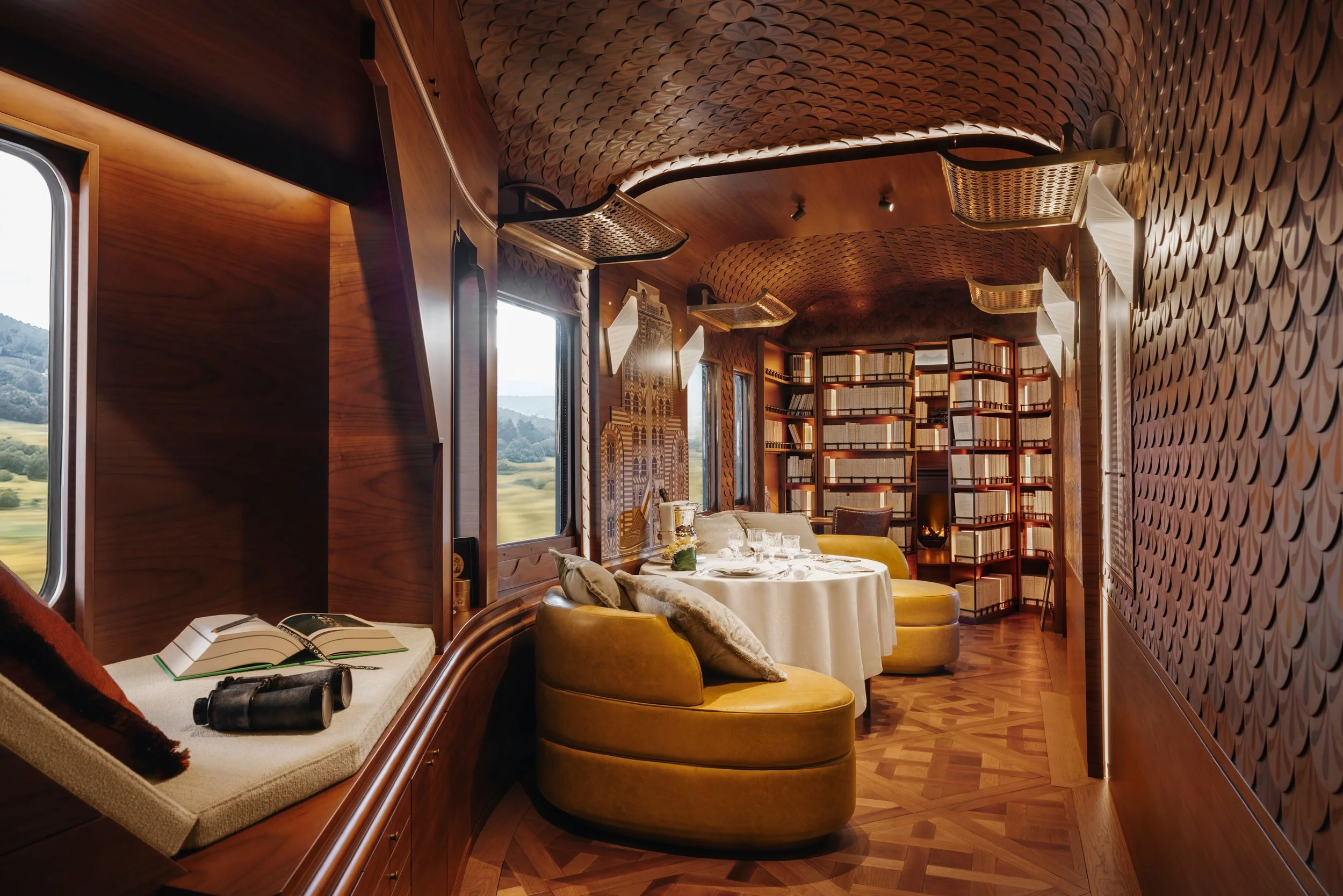 A cozy interior room with wooden walls, a table set for dining with cloth, glassware, and candles, surrounded by plush yellow chairs, and decorated with bookshelves and scenic window views.