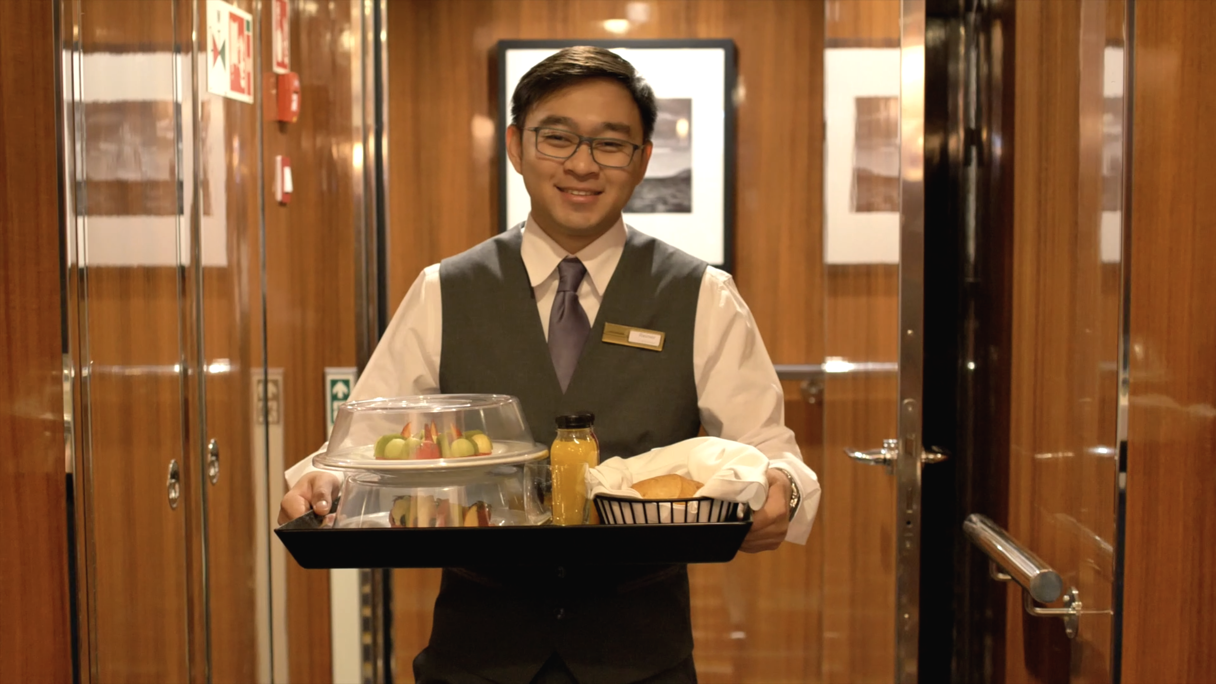 A smiling hotel employee or waiter dressed in a white shirt, tie, and vest carrying a tray with food and drinks inside an elevator with wood paneling and framed pictures on the wall.