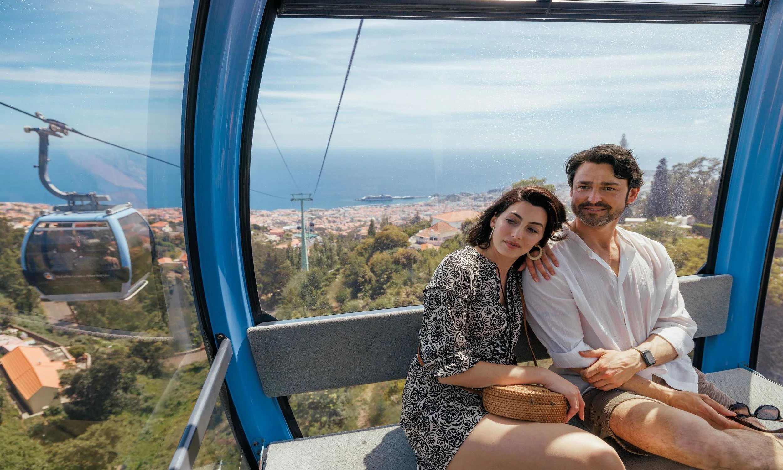 A couple sitting inside a blue gondola on a scenic mountainside, with a view of a town, trees, and the ocean in the background.