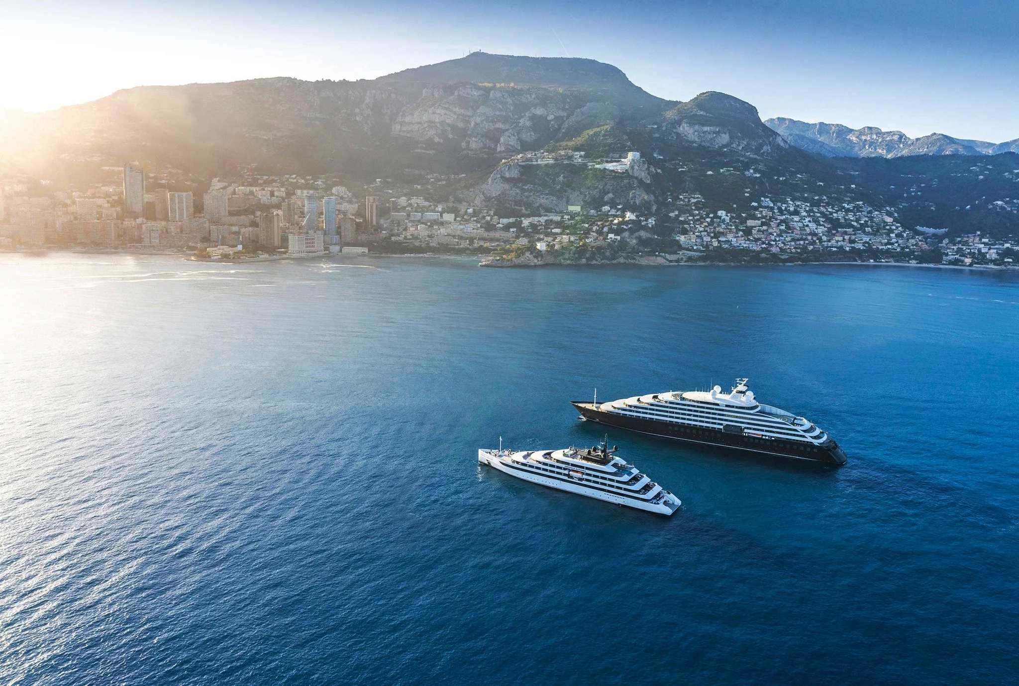 Aerial view of Monaco coastline with high-rise buildings, mountains in the background, and two large luxury yachts sailing on the water.