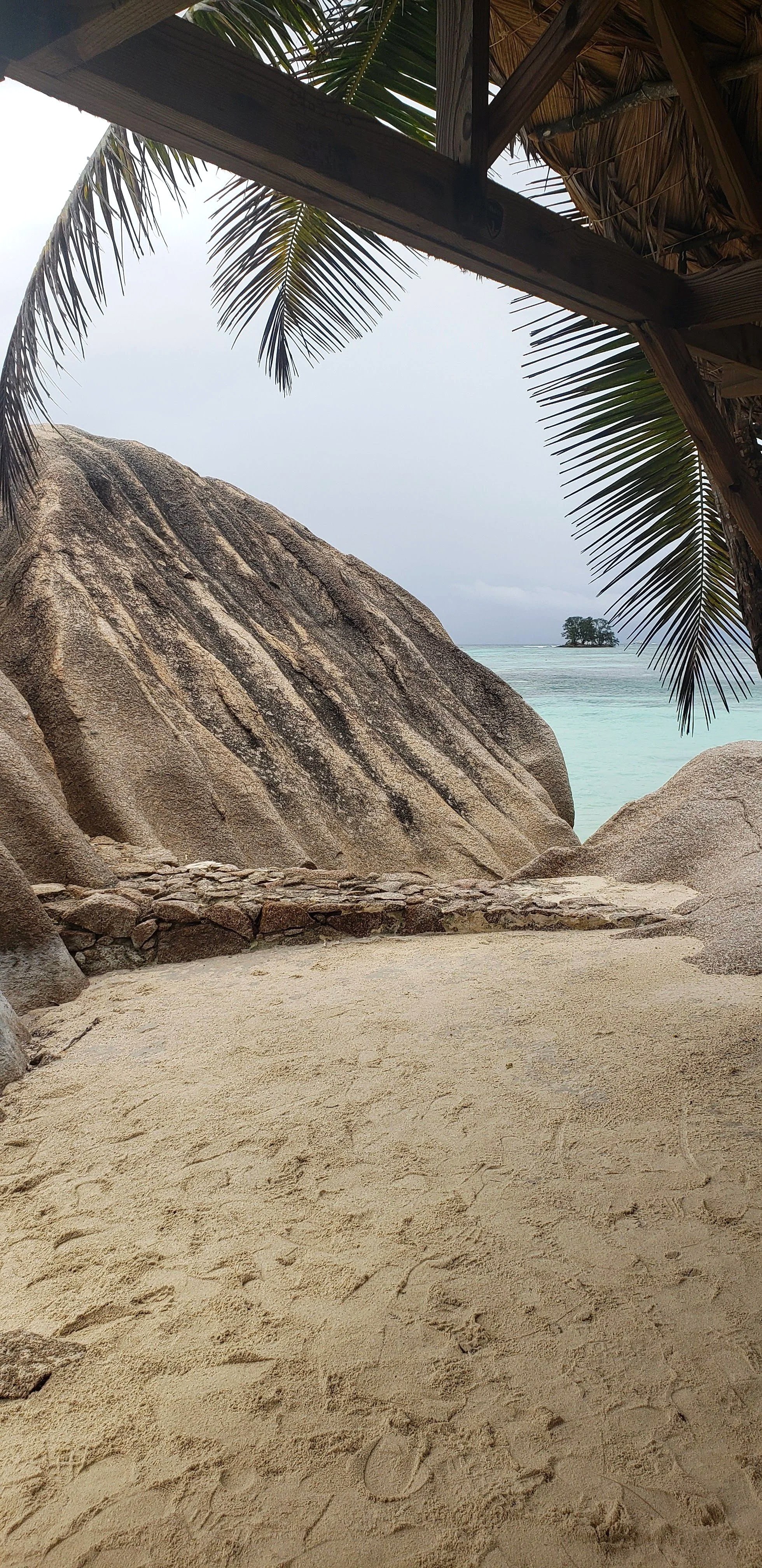 Tropical beach scene with sandy ground, large granite rocks, palm tree leaves overhead, and a small island in the distance over calm turquoise waters.