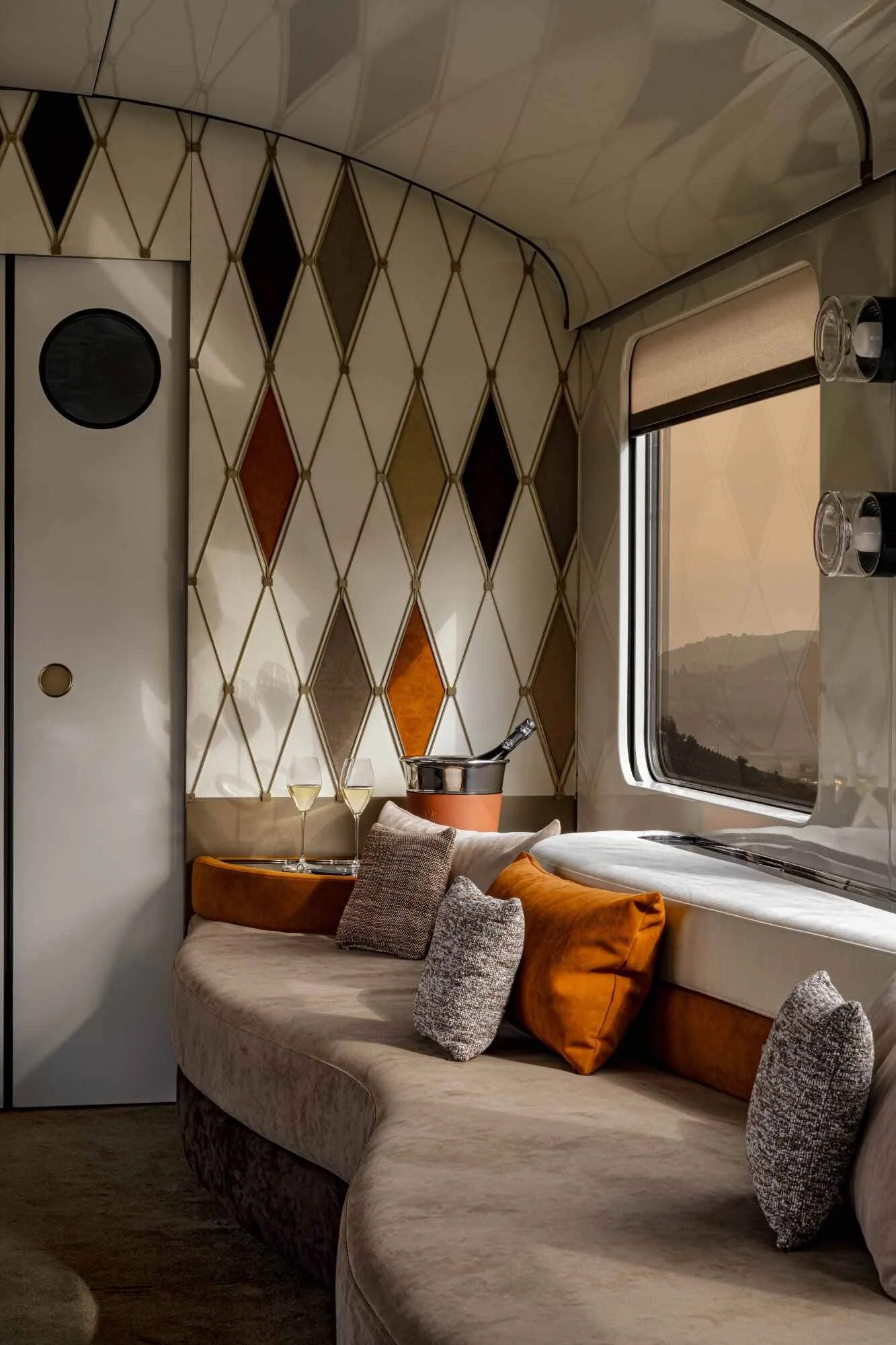 Interior of a cozy train compartment with a curved beige sofa adorned with gray and rust-orange pillows, a small wooden table next to the sofa holding two glasses of white wine, a window with a beige blind, and a distinctive diamond-patterned wall with various shades of brown and beige.