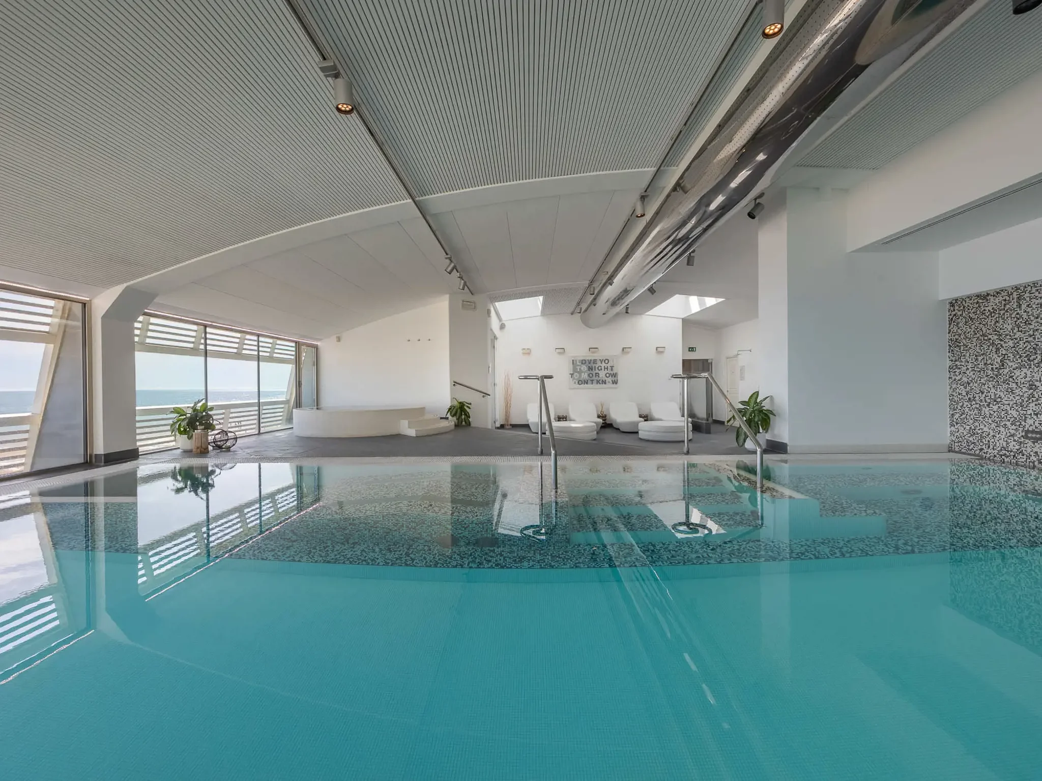 Indoor swimming pool with a tiled floor, chairs, plants, and large windows showing an ocean view.