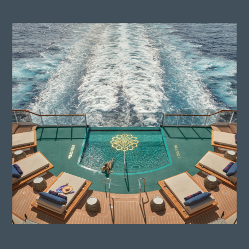 View of the back of a cruise ship showing a swimming pool surrounded by lounge chairs and towels, with the sea and the ship's wake in the background.
