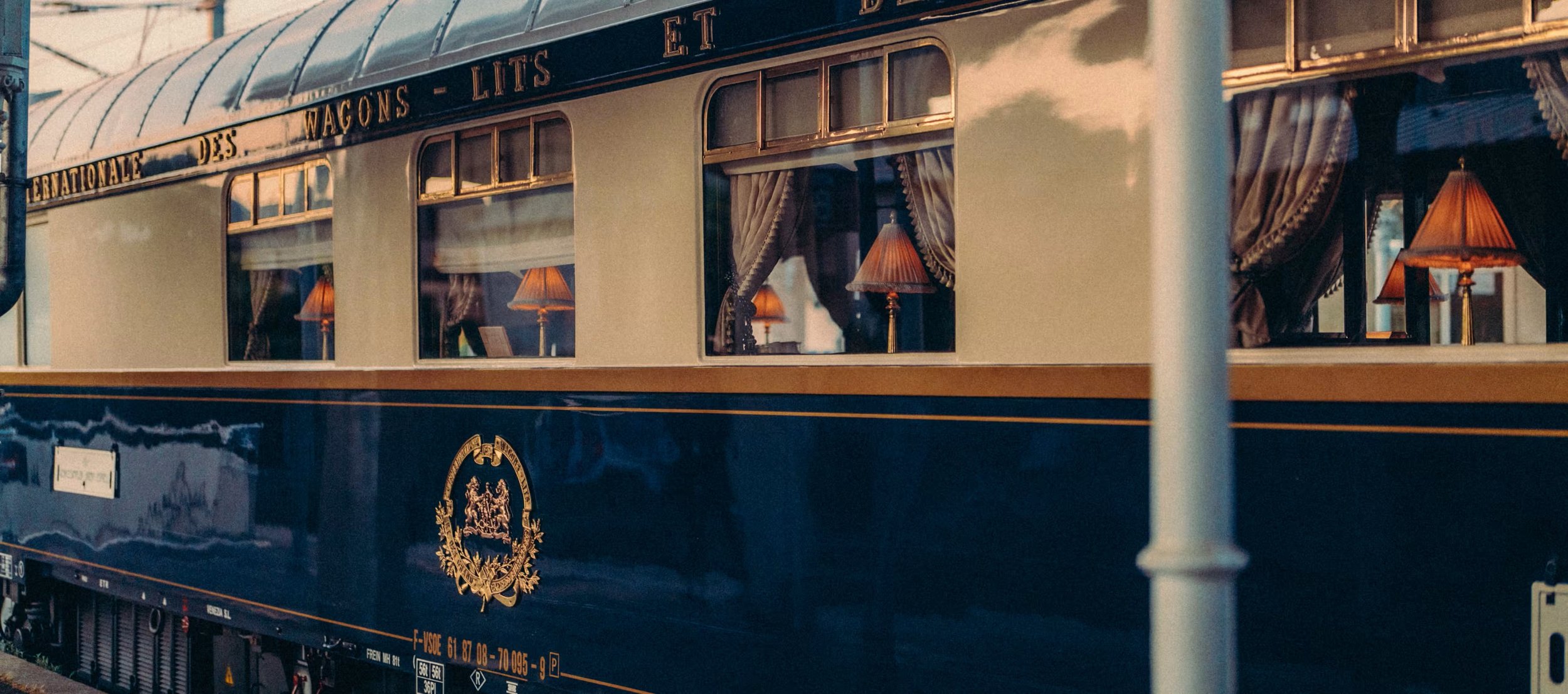 A vintage luxury train car with navy blue and gold exterior, featuring large windows with curtains and lamps inside.
