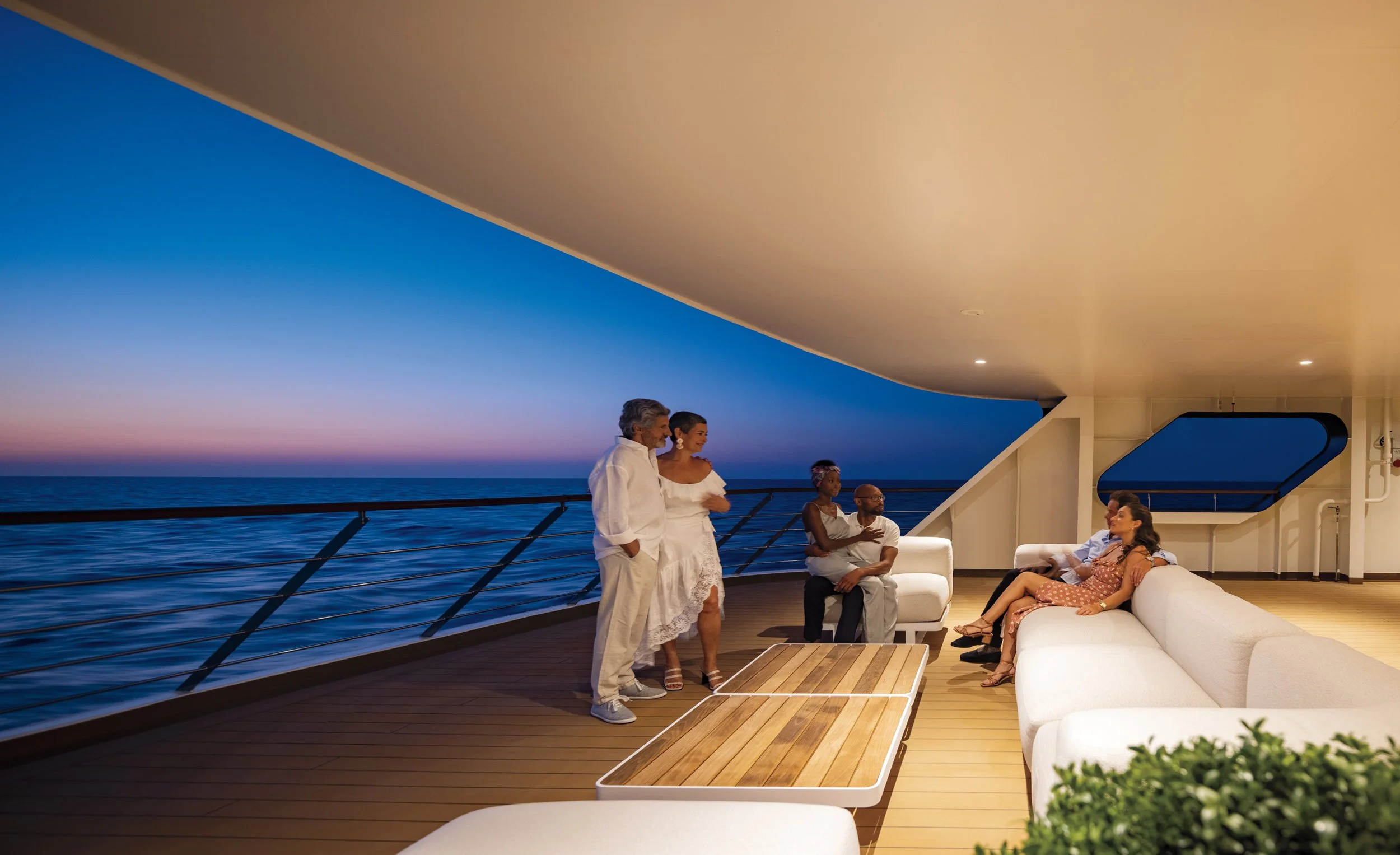 A group of people socializing on a yacht deck during sunset, with the ocean and colorful sky in the background.