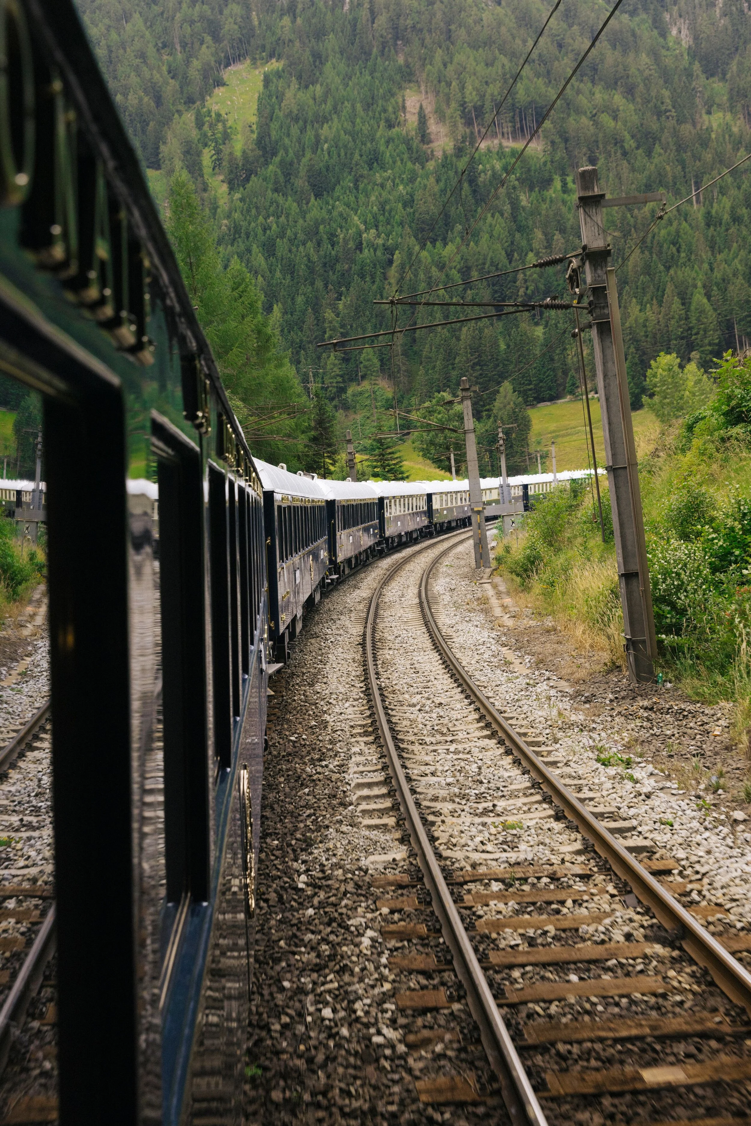 A train moving along curved railway tracks through a mountainous, forested landscape with green trees and hills in the background, under power lines.