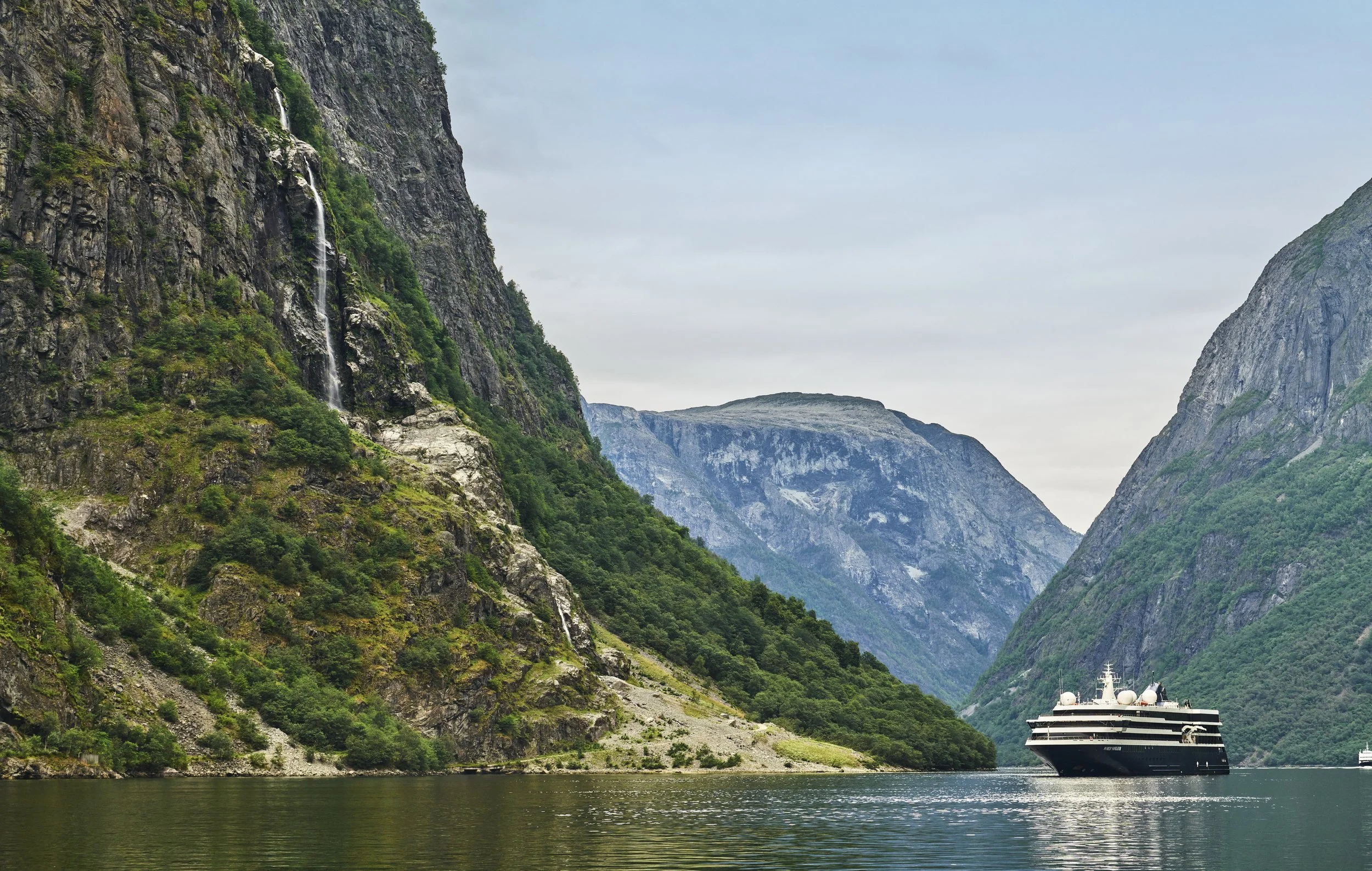 Cruise ship sailing through a deep fjord surrounded by steep, rugged mountains covered in greenery, with waterfalls cascading down the cliffs on the left side and cloudy skies overhead.