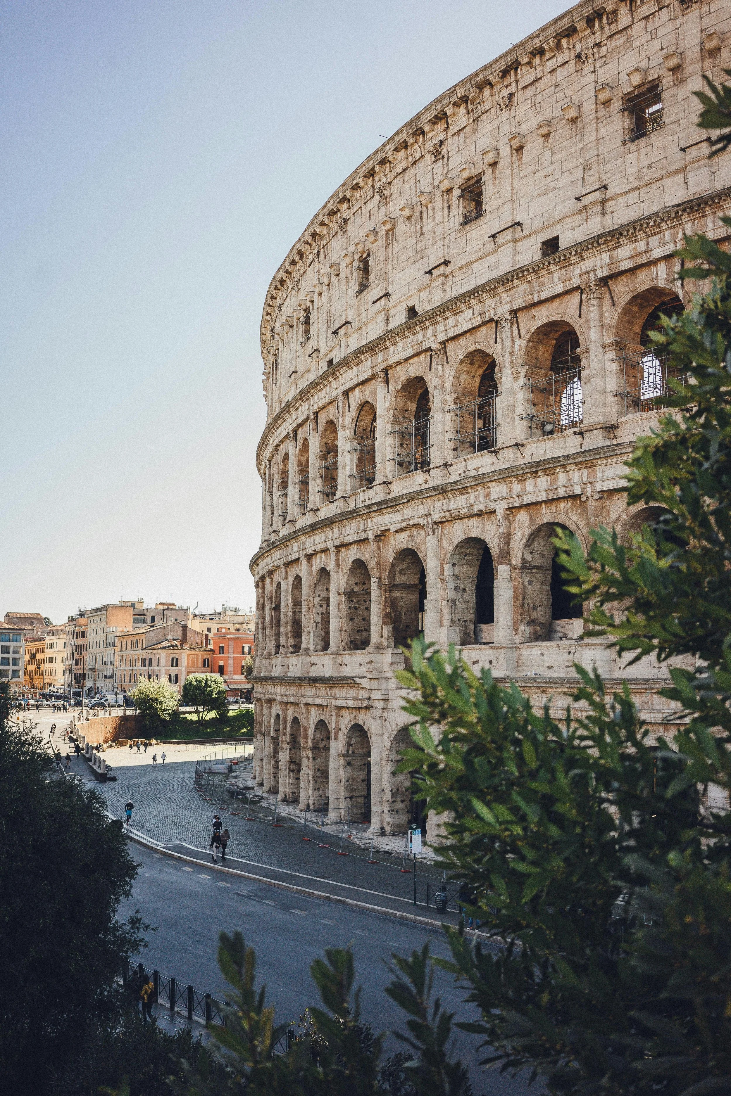 The image shows the Colosseum in Rome, Italy, a large ancient amphitheater with multiple arches and a partially open upper level. The sky is clear, and there are people walking around the area, with some trees and buildings in the background.