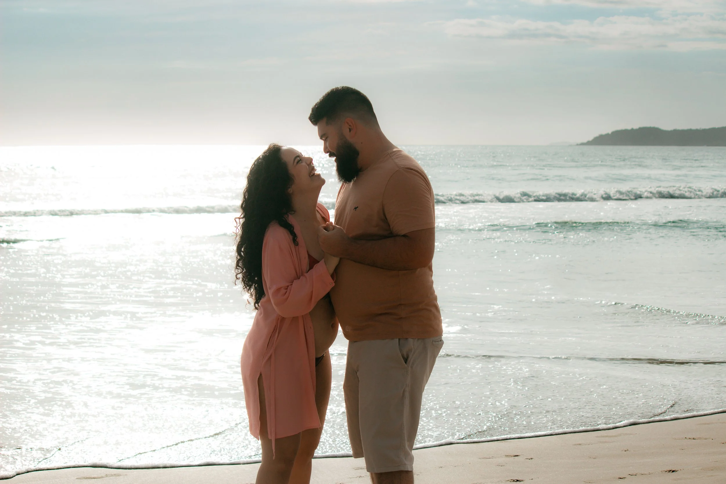 A couple smiling and embracing on the beach during sunset with ocean waves and a distant landmass in the background.
