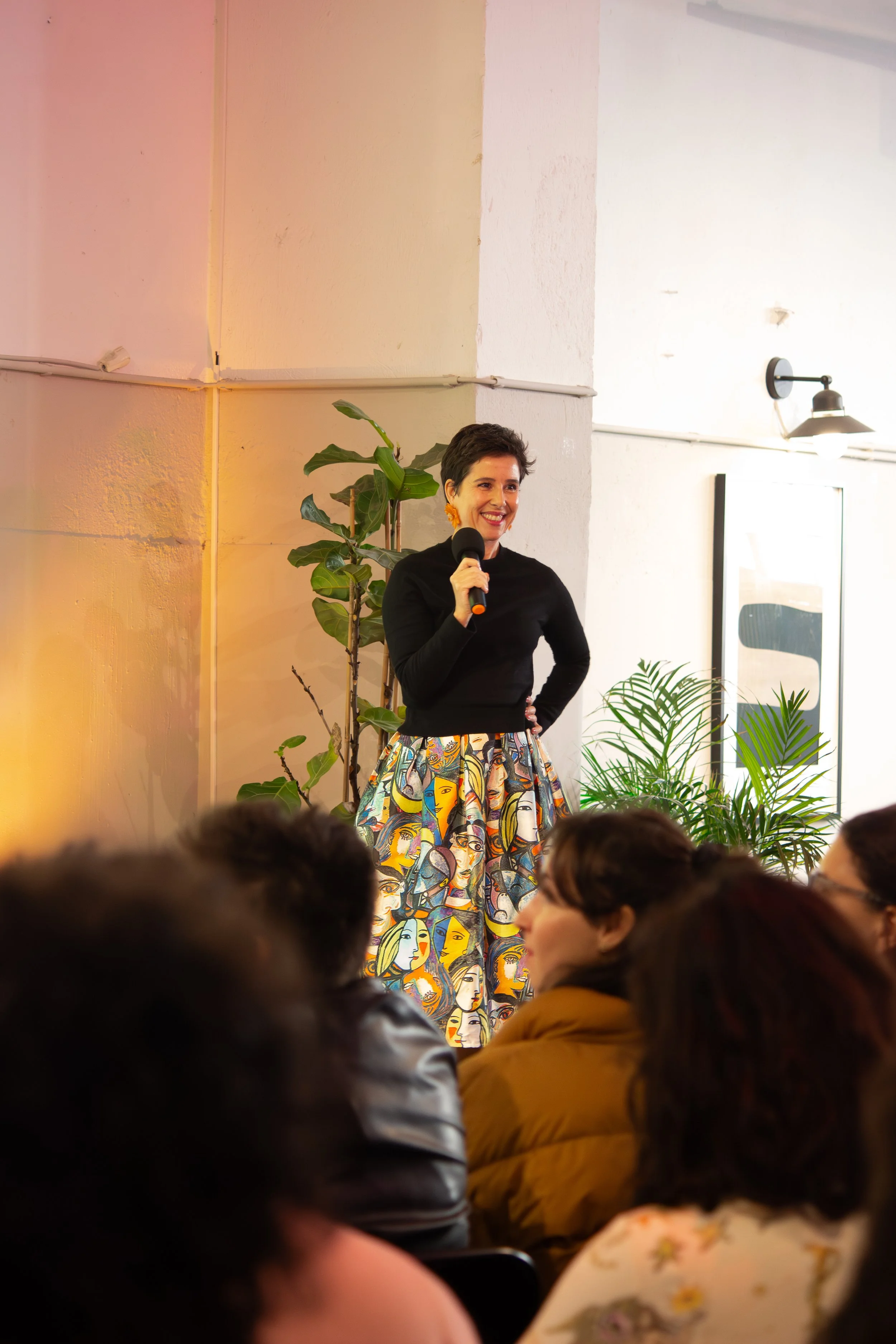 A woman holding a microphone and smiling, standing in front of a white wall with plants, surrounded by an audience.