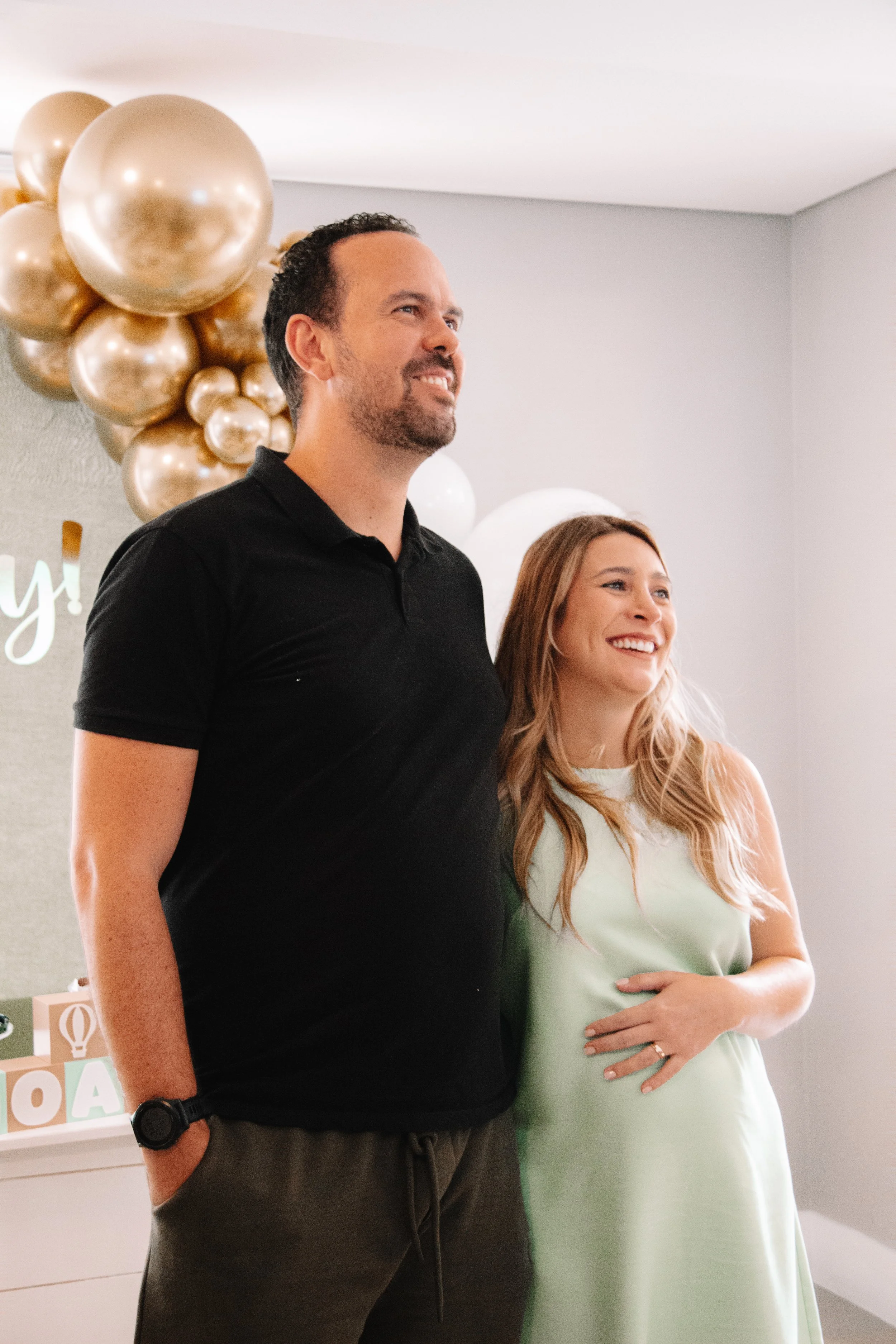 A man and a pregnant woman standing close together at a celebration, smiling. The man has short curly hair and a beard, wearing a black polo shirt with a watch on his wrist. The woman has long, wavy blonde hair, wearing a light green dress and an engagement ring on her left hand, which she touches her baby bump with. Behind them, there are gold and white balloons and a light-colored wall with part of a sign visible.