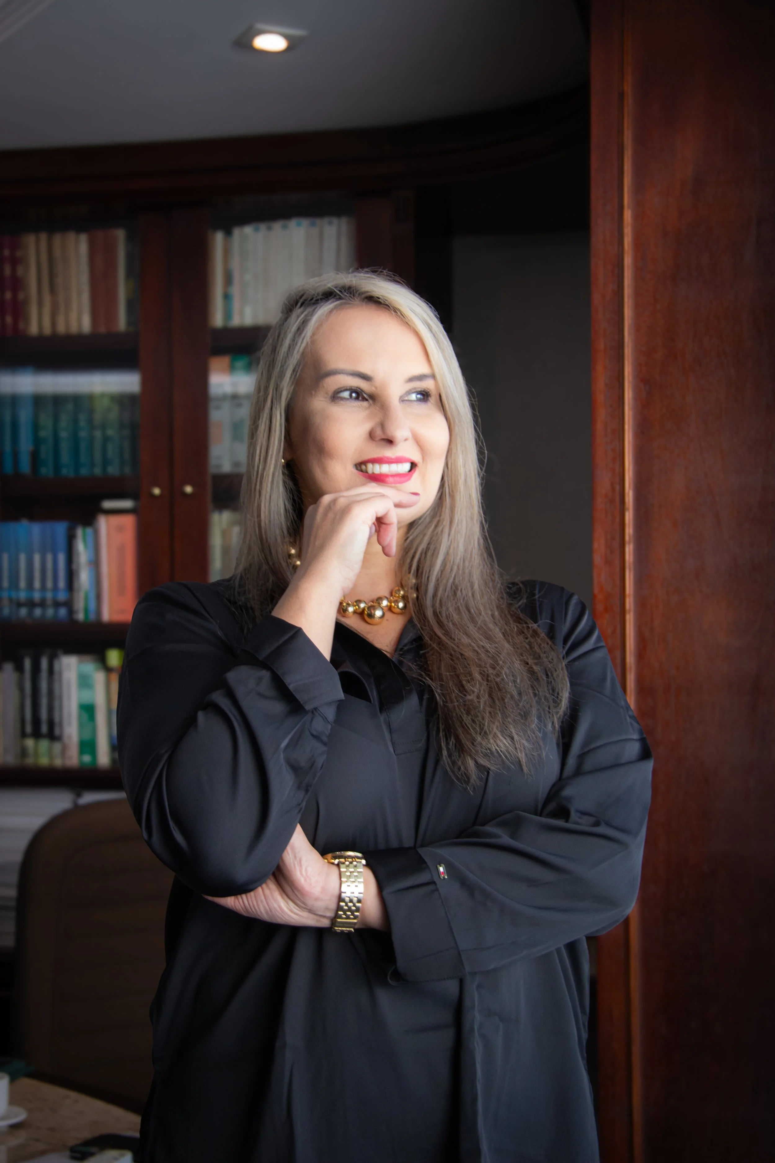 A woman with long blonde hair, wearing a black shirt, gold necklace, and watch, stands in front of a bookcase with her hand resting on her chin, looking to her left with a smile.