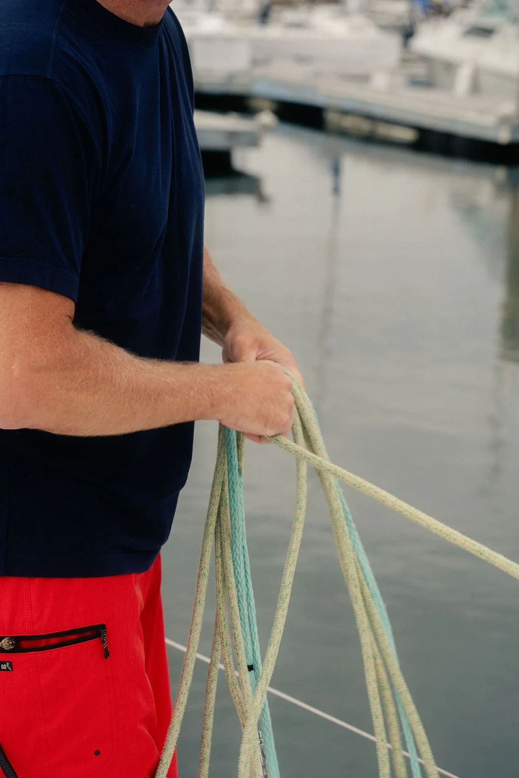 A man in a navy blue shirt and red shorts handling several ropes on a dock with boats in the background.