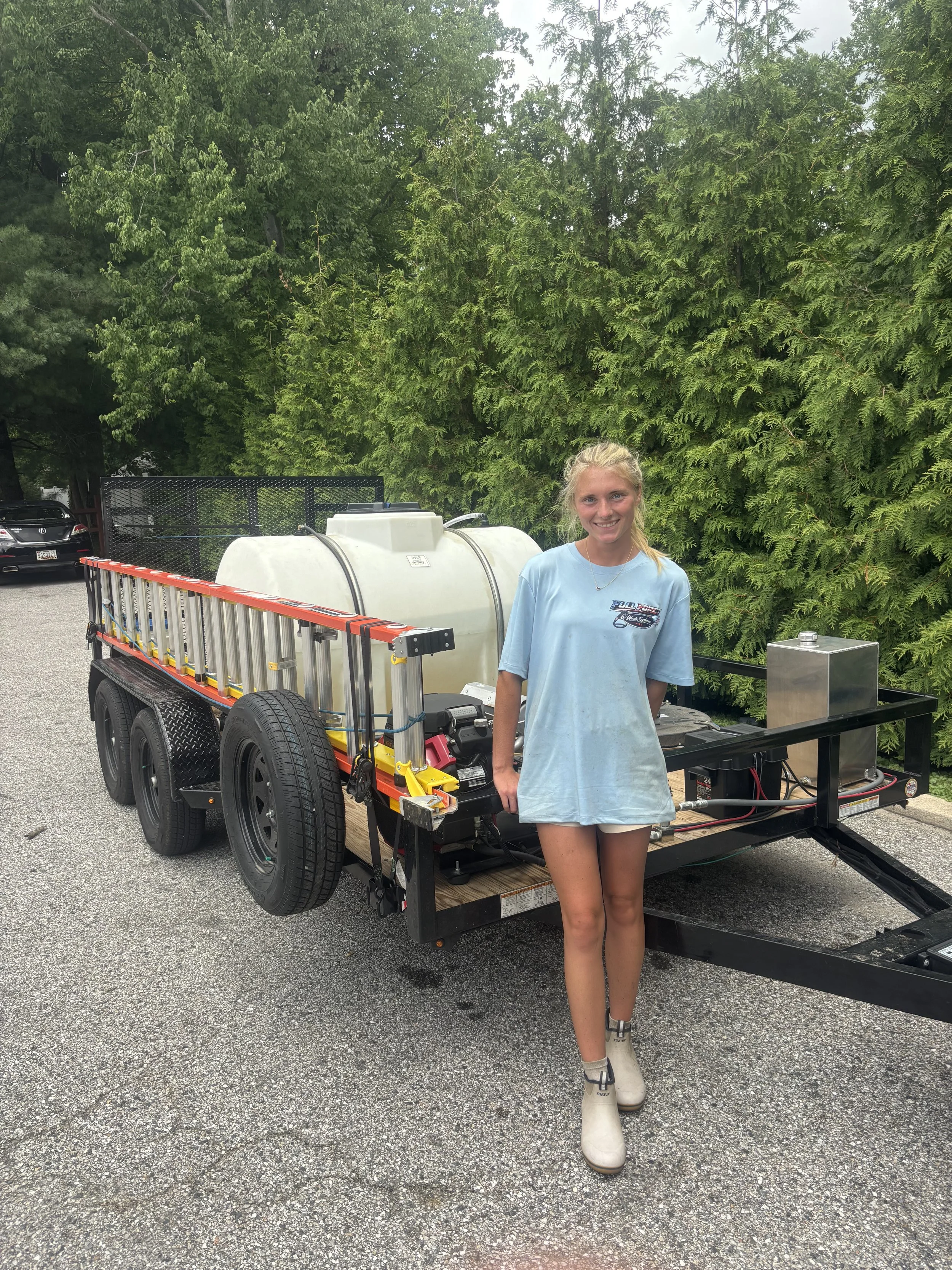 A young woman with blonde hair, wearing a light blue T-shirt and beige boots, standing outdoors next to a utility trailer with equipment, with trees in the background.