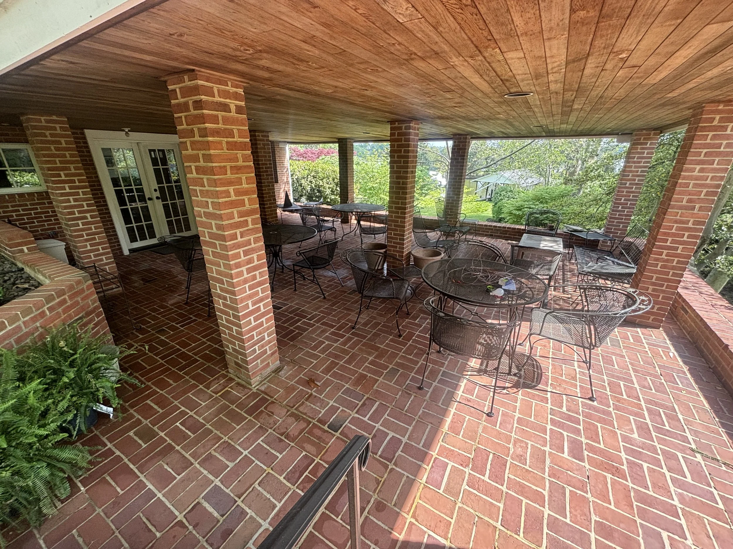 Covered brick patio with black metal outdoor furniture, surrounded by green trees and bushes.