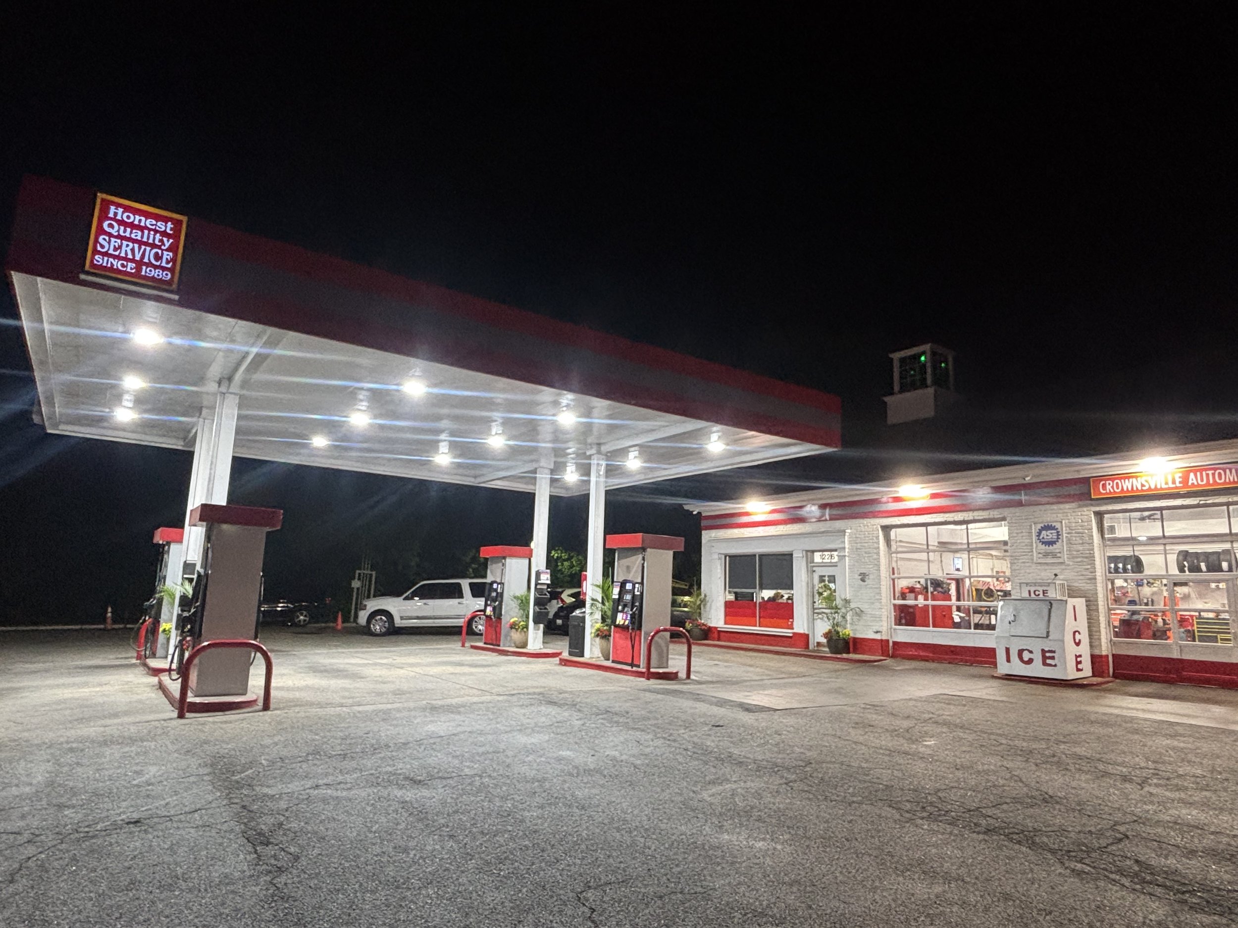 Night view of a Crownsville AutoMall gas station with a sign showing ''Honest Quality Service Since 1989.'' The station is well-lit, with parked cars and an ice vending machine visible.