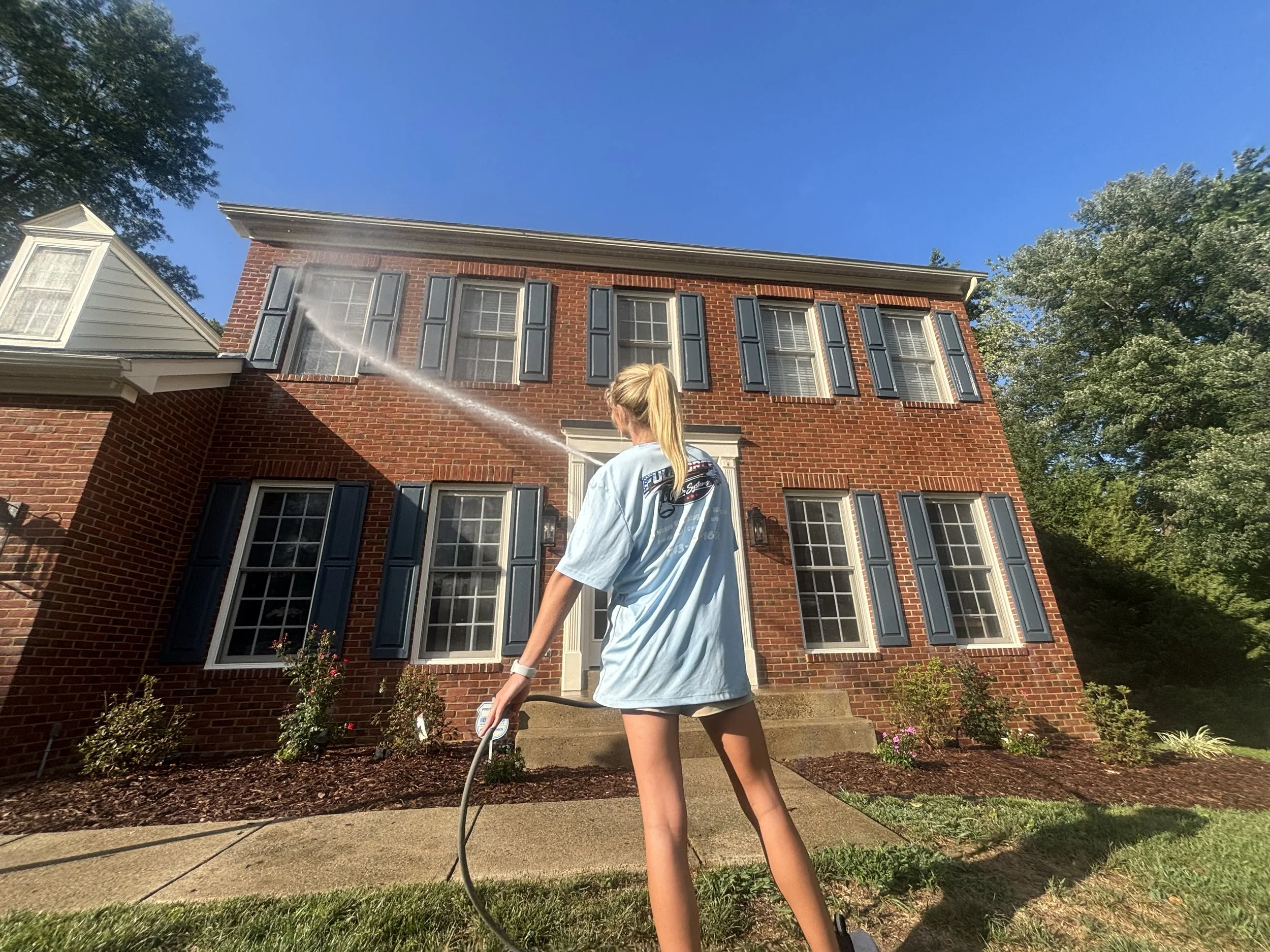 A woman watering flowers in front of a three-story brick house with four windows and black shutters, under a clear blue sky, with a large tree on the right side.
