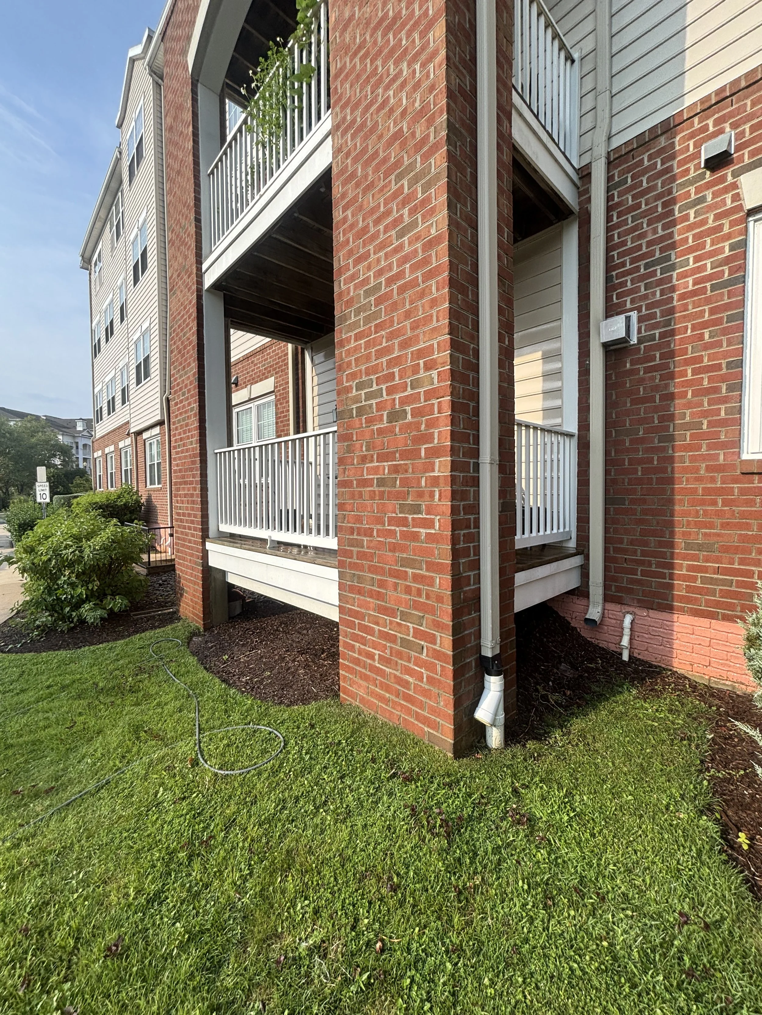 View of the exterior of a brick apartment building with green grass, a garden hose, and shrubbery in the foreground. The building has small balconies with white railings, and visible drainage pipes run along the wall.