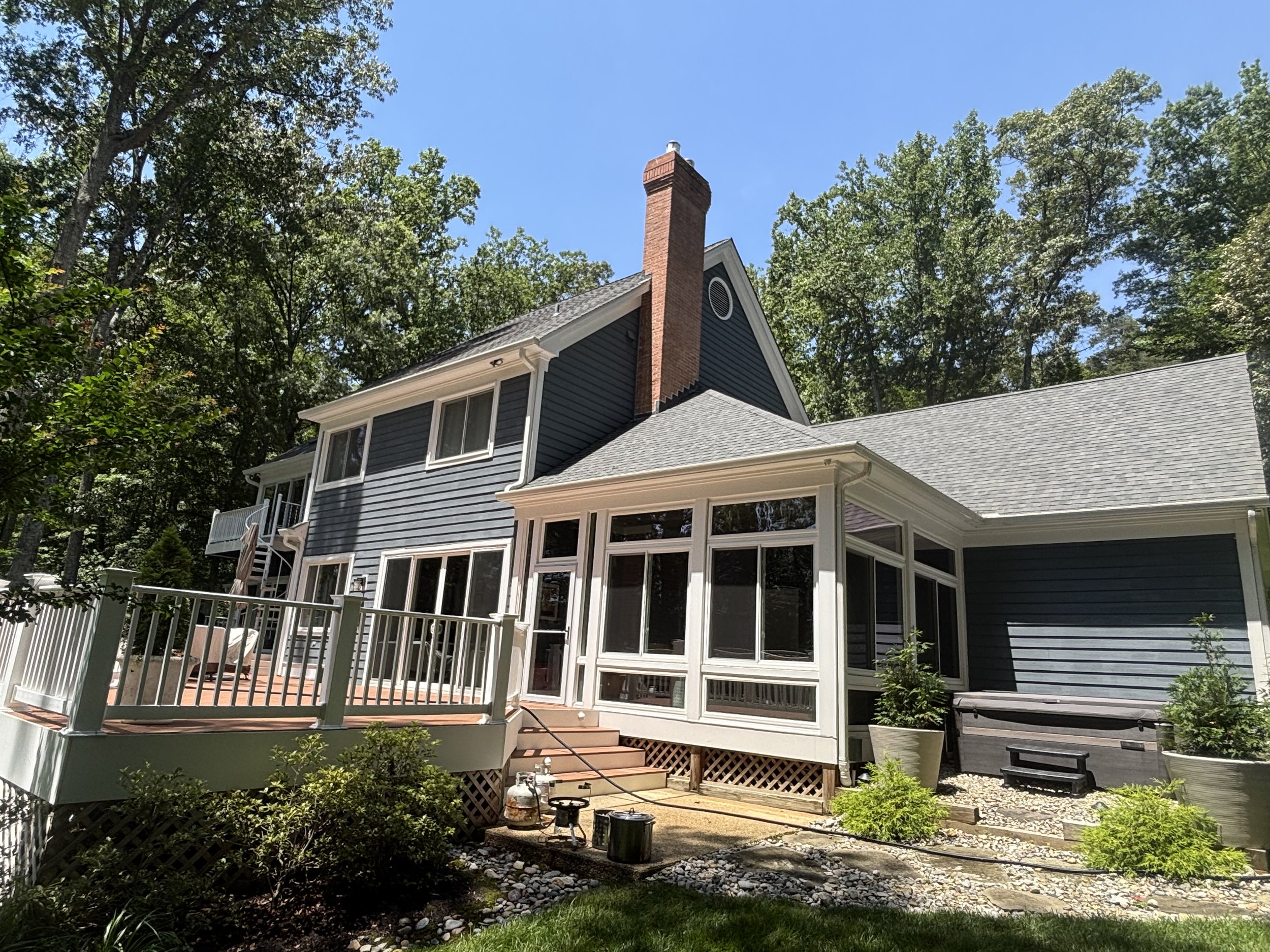 Rear view of a blue house with a deck, surrounded by trees and greenery, under a clear blue sky.