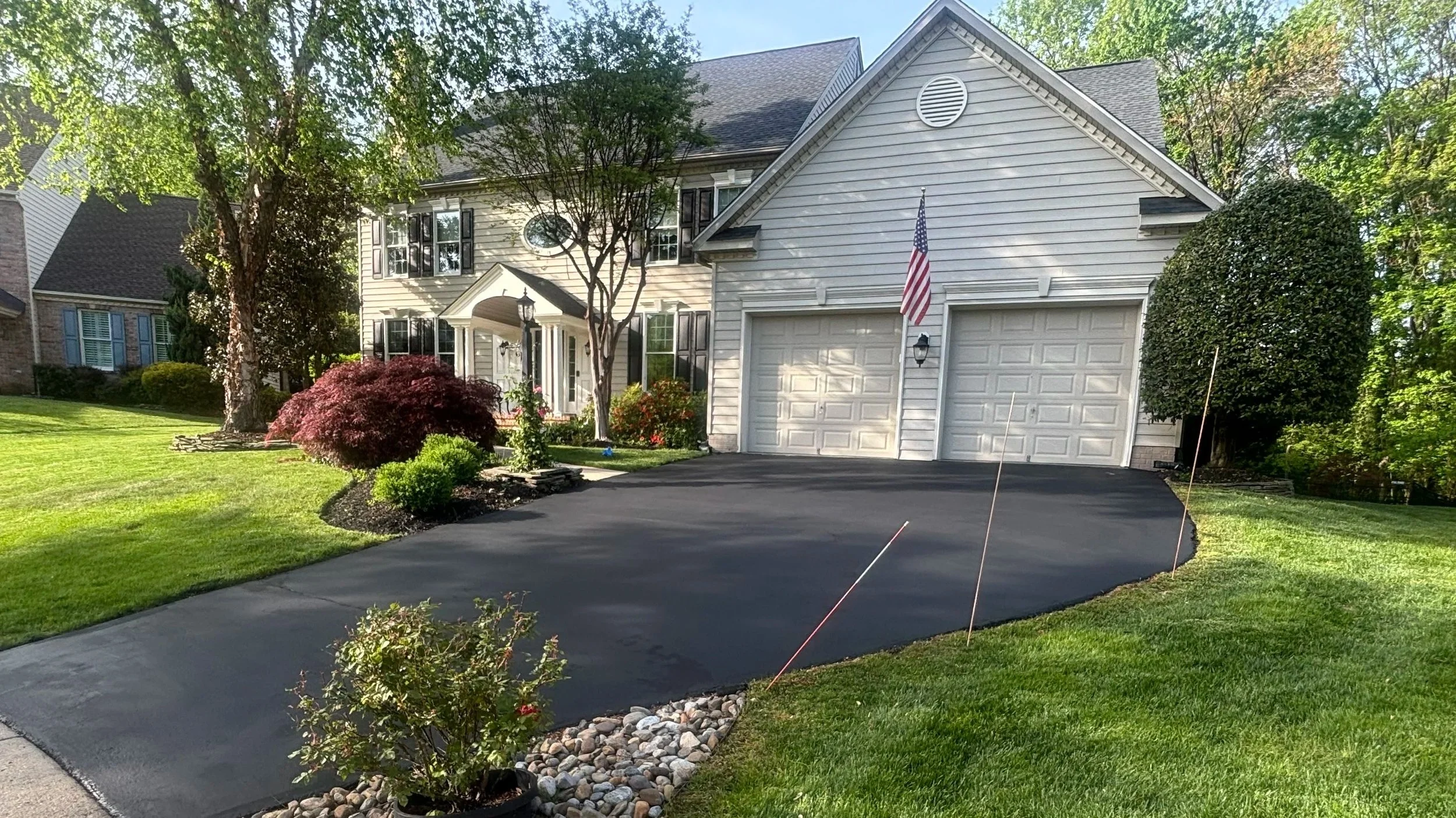 Front view of a suburban house with white siding, a two-car garage, and a well-maintained lawn with trees and landscaped bushes.