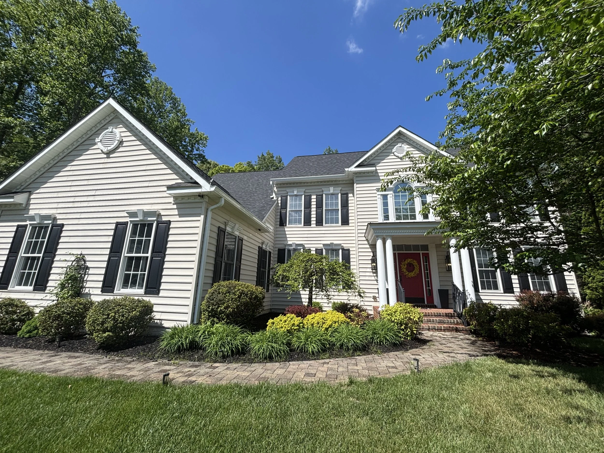 A white two-story house with black shutters, a red front door with a yellow wreath, surrounded by green bushes and plants, under a clear blue sky with some clouds.
