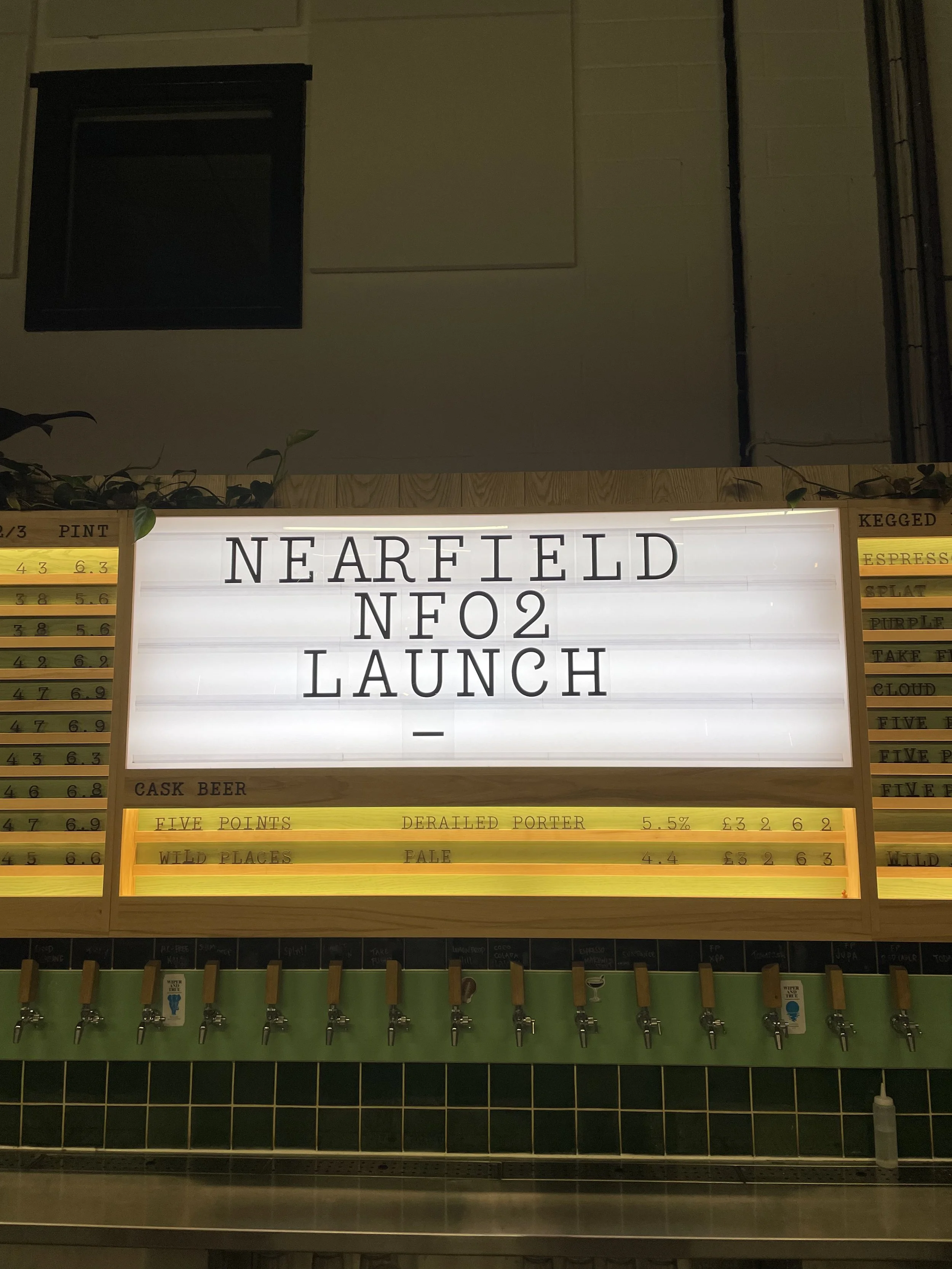 A brightly lit menu board with the words 'NEARFIELD NE02 LAUNCH' on it, flanked by price and drink options, and beer taps below.