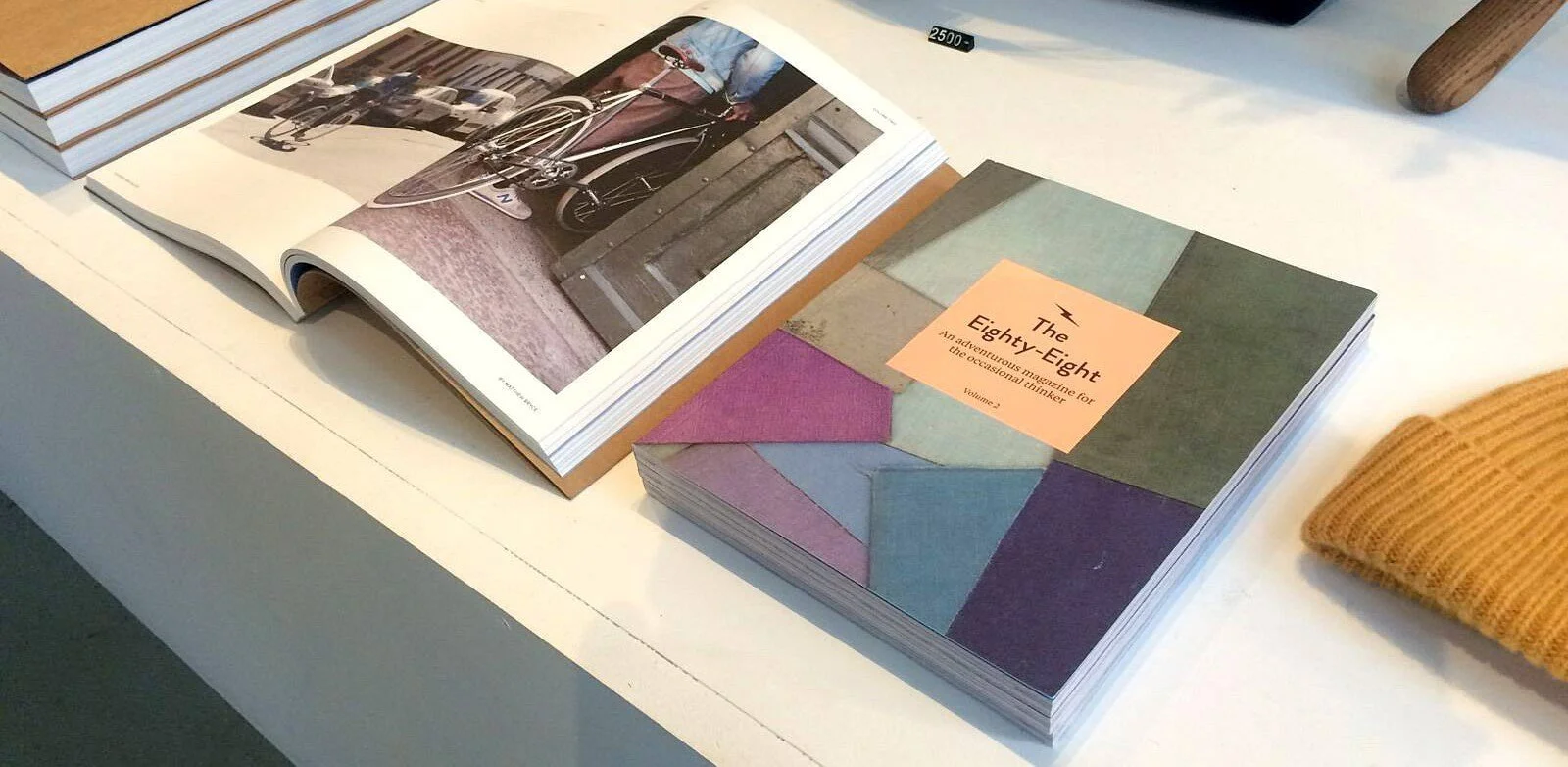 Open magazine featuring photographs of a bicycle and street scene, beside a closed magazine titled "The Eighty-Eight" with colorful geometric cover design on a white table.