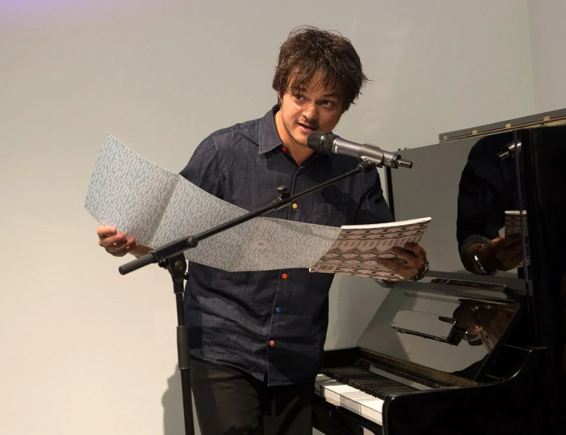 A man singing or speaking into a microphone while holding sheet music or lyrics, standing next to a piano, on a stage or in a performance setting.