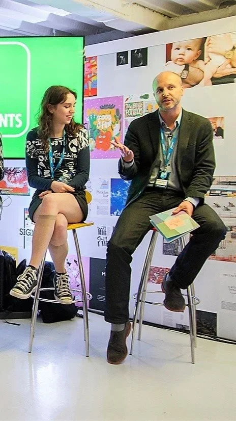 A man and a woman seated on bar stools in conversation at an event. The woman is wearing a black and white jacket, black skirt, and sneakers, while the man is dressed in a navy blazer, blue shirt, and black pants, holding a booklet. They are in front of a colorful backdrop with posters and photos, including a baby.