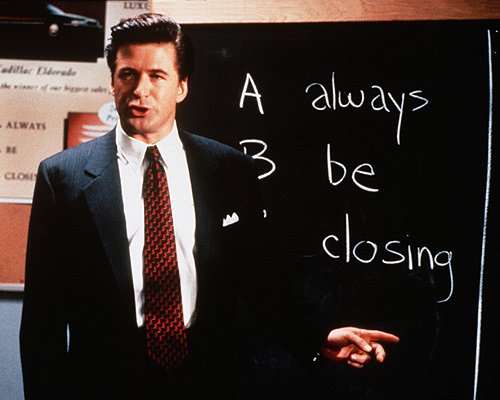 The Alec Baldwin character from Glengarry Glen Ross in a suit standing in front of a blackboard with writing that says, 'A always B, B be closing.'