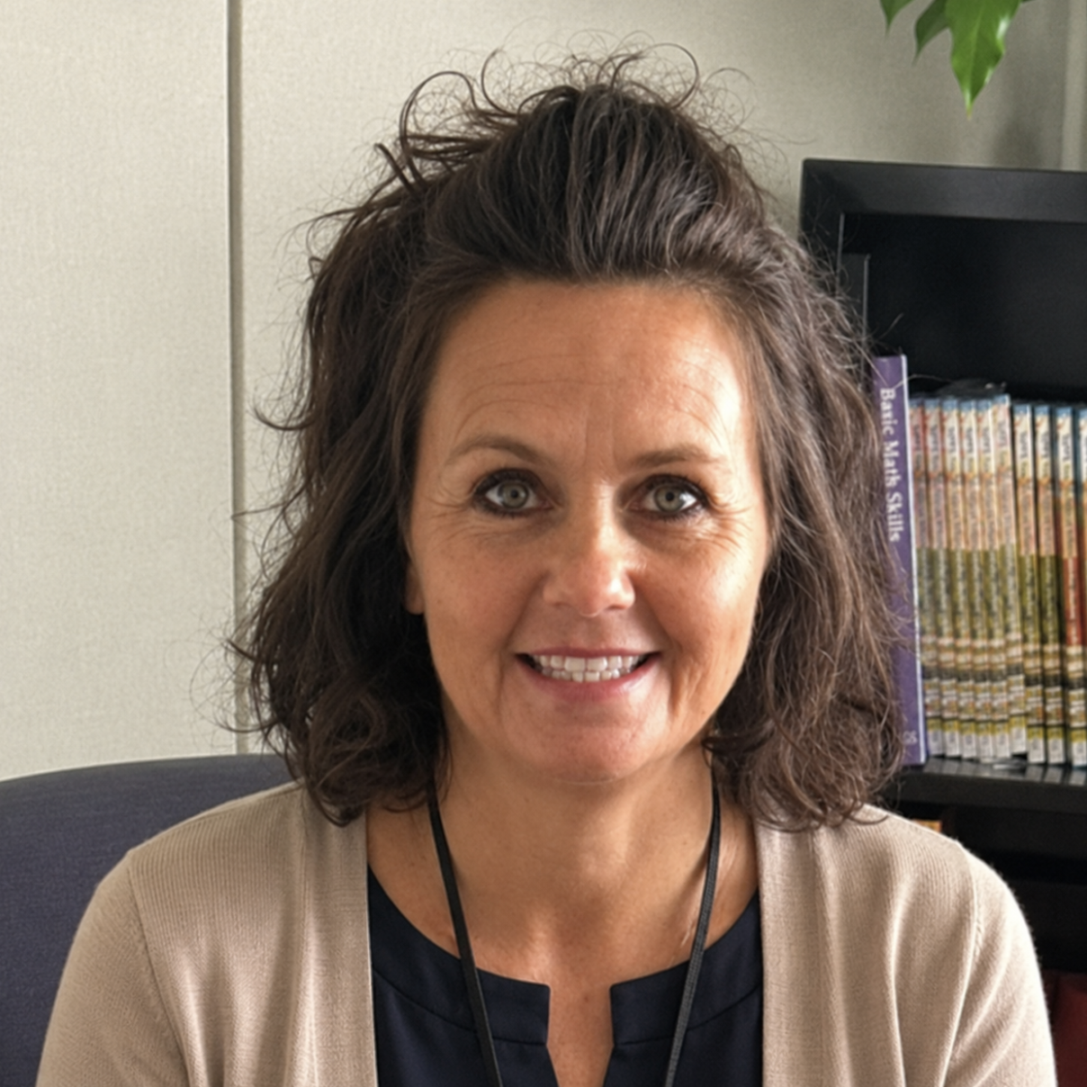 A woman with shoulder-length wavy brown hair smiling, sitting in an office with a bookshelf of books behind her.