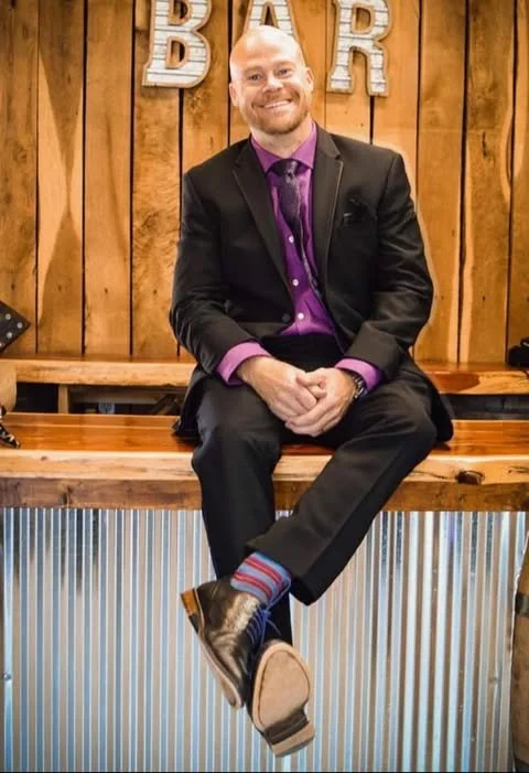 Man dressed in a black suit with a purple shirt and tie, sitting on a wooden bench in front of a wood-paneled wall with the word 'BAR' displayed behind him, smiling with his hands clasped, wearing colorful striped socks, and stylish shoes.