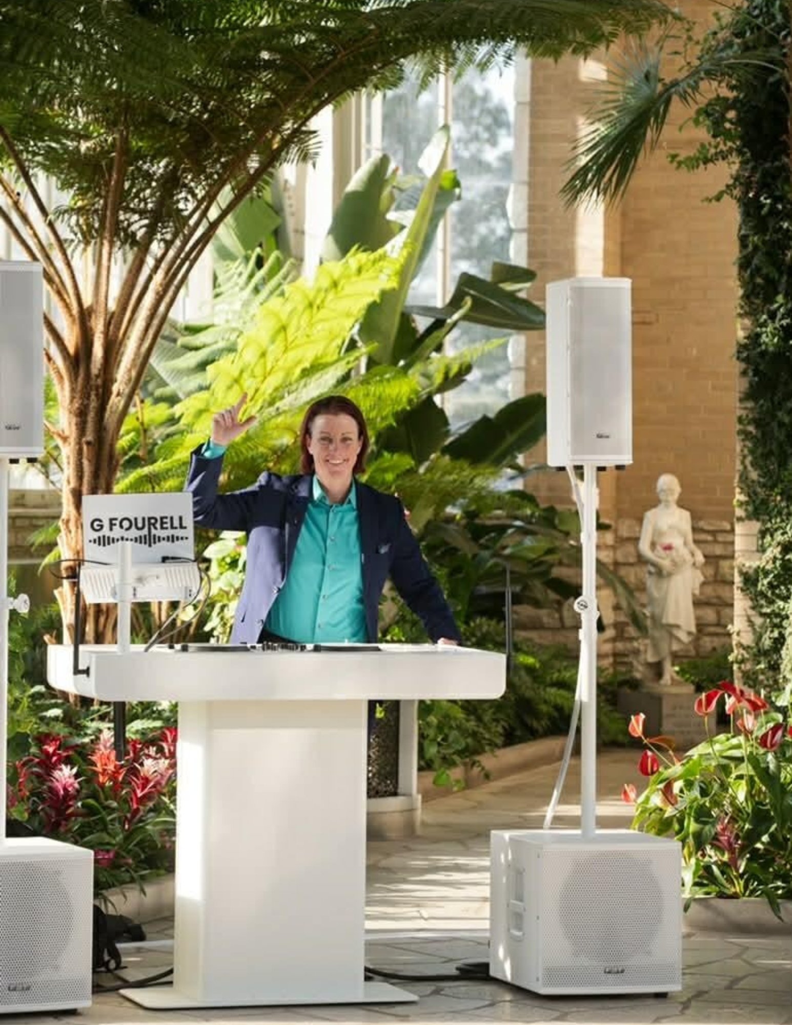 A woman standing on a white elevated platform in a greenhouse or botanical garden. She is smiling and waving, dressed in a dark blazer and teal shirt. There are large green plants and flowers around her, with a bust sculpture visible in the background. The scene appears to be set for an event or presentation, with speakers and audio equipment present.