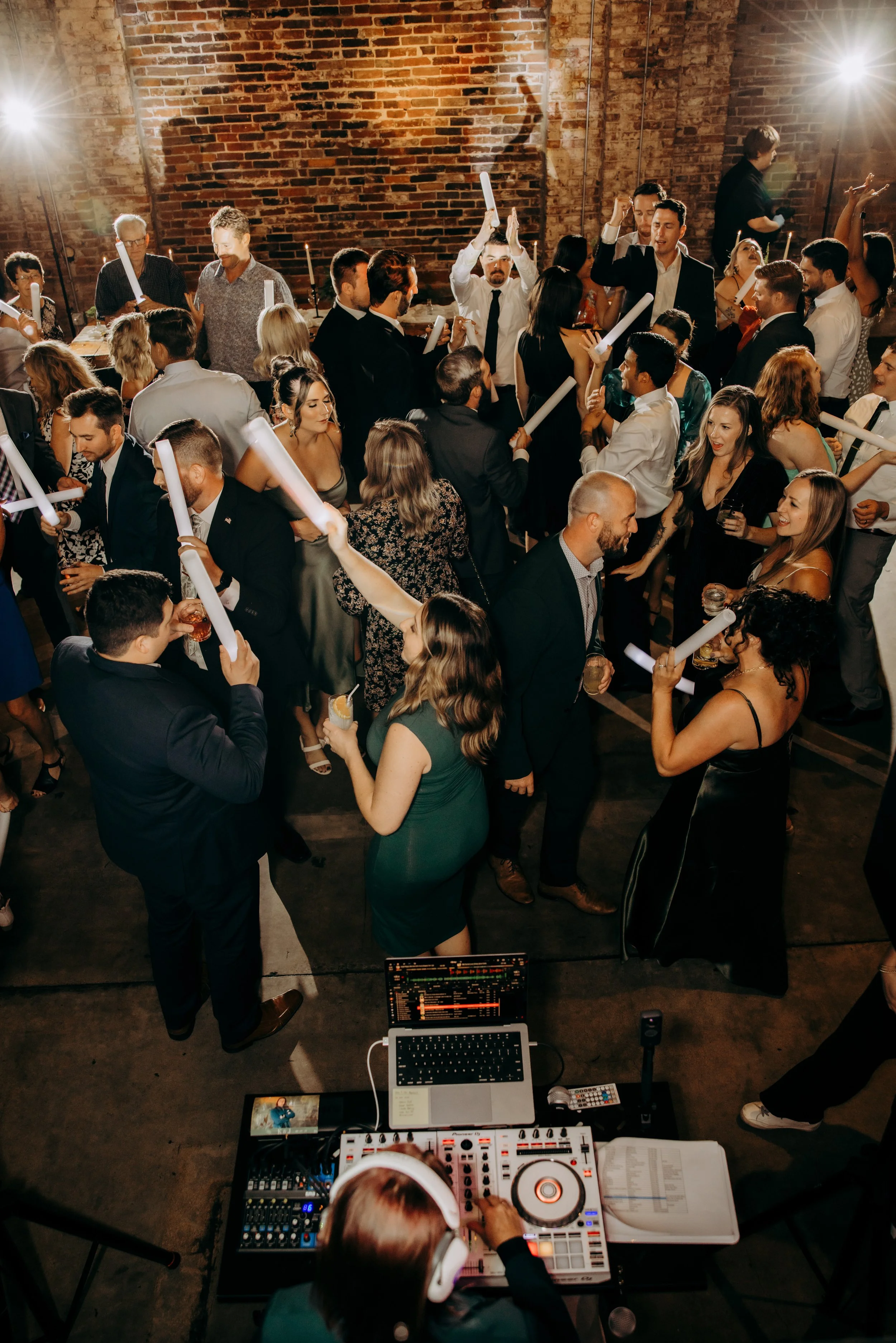 A crowded dance party with people dancing and socializing in front of a brick wall. A DJ is seen from behind at a mixing console, playing music for the crowd.