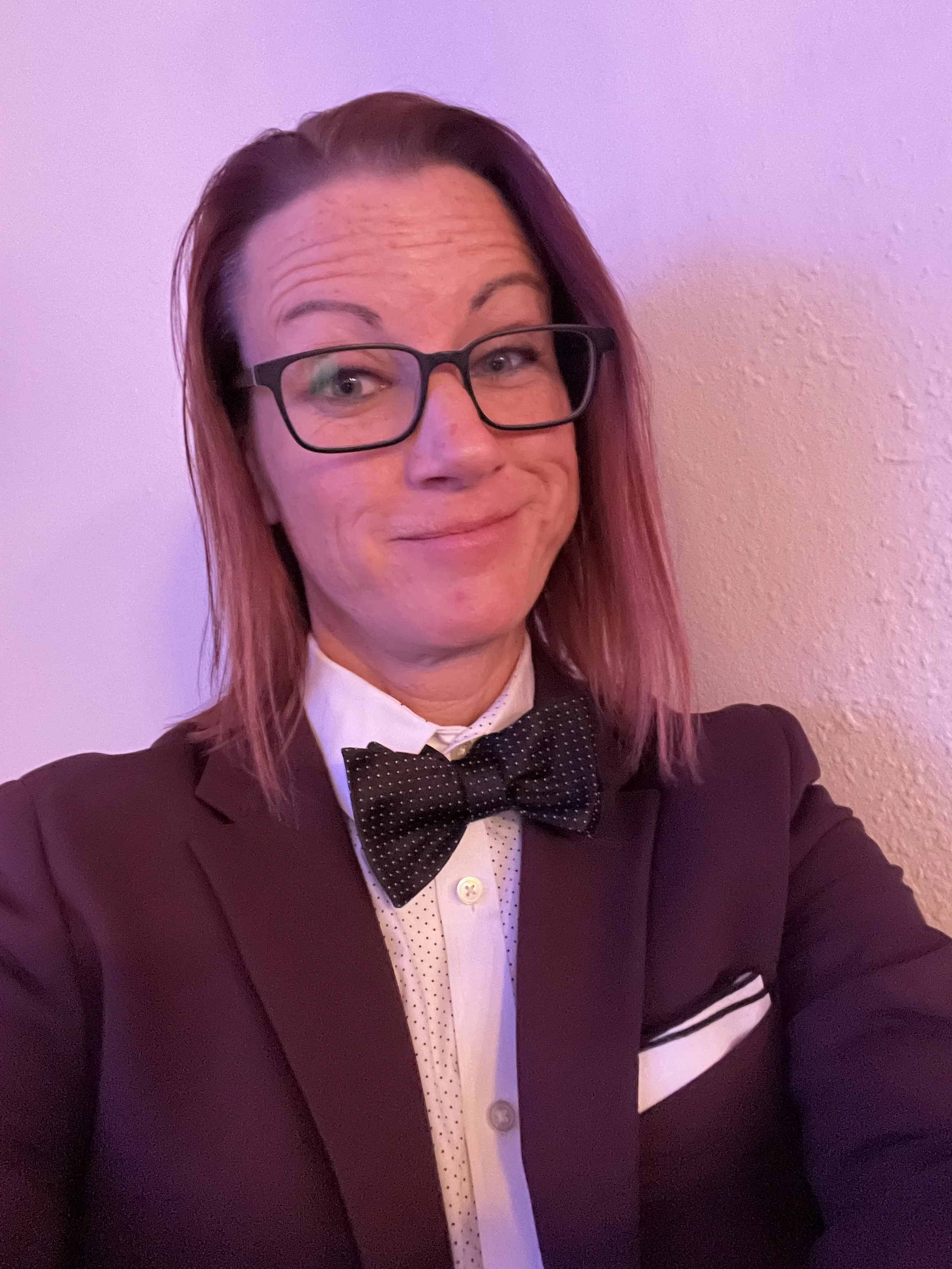 A person with shoulder-length pink hair, glasses, smiling, wearing a black tuxedo with a white shirt and black polka dot bow tie, posing against a textured pink wall.