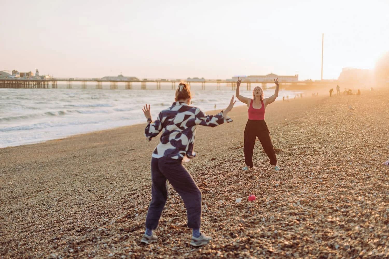 Two women on a beach playing with a ball during sunset, with a pier in the background.